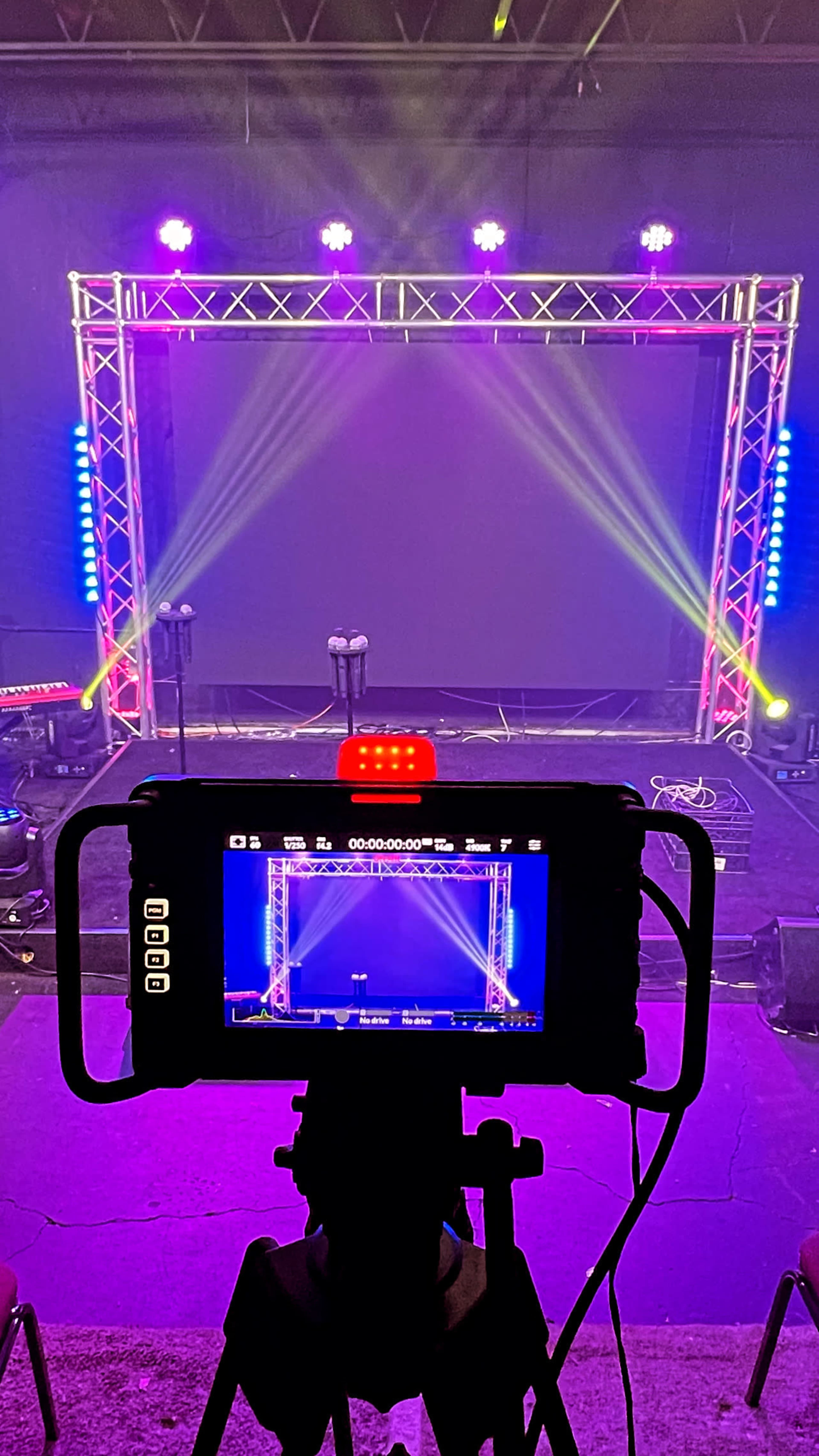 A camera on a tripod aimed at a stage setup with colorful lights and a large screen in the background.