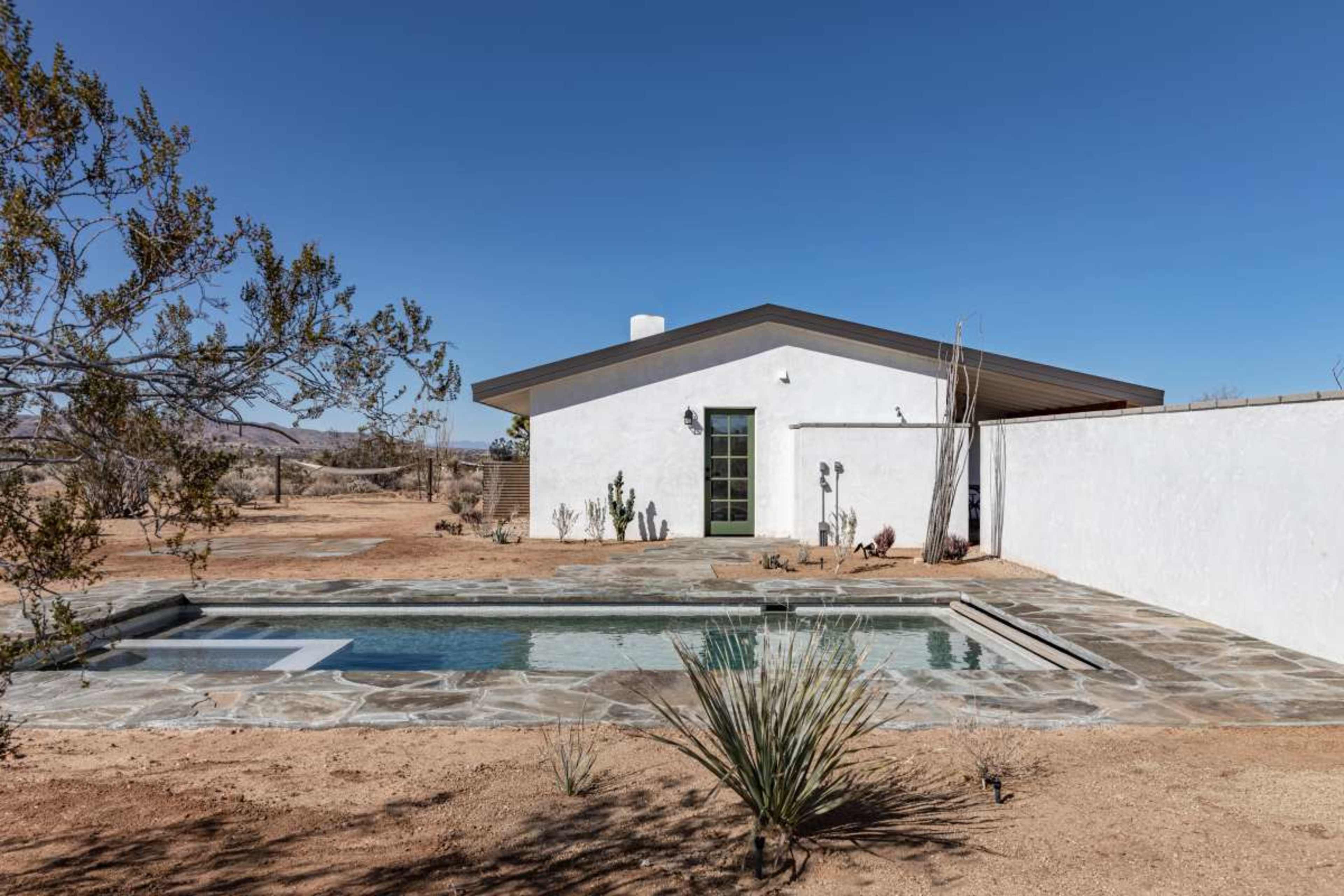 A modern white building with a flat roof is set beside a pool in a desert landscape, surrounded by sparse vegetation.