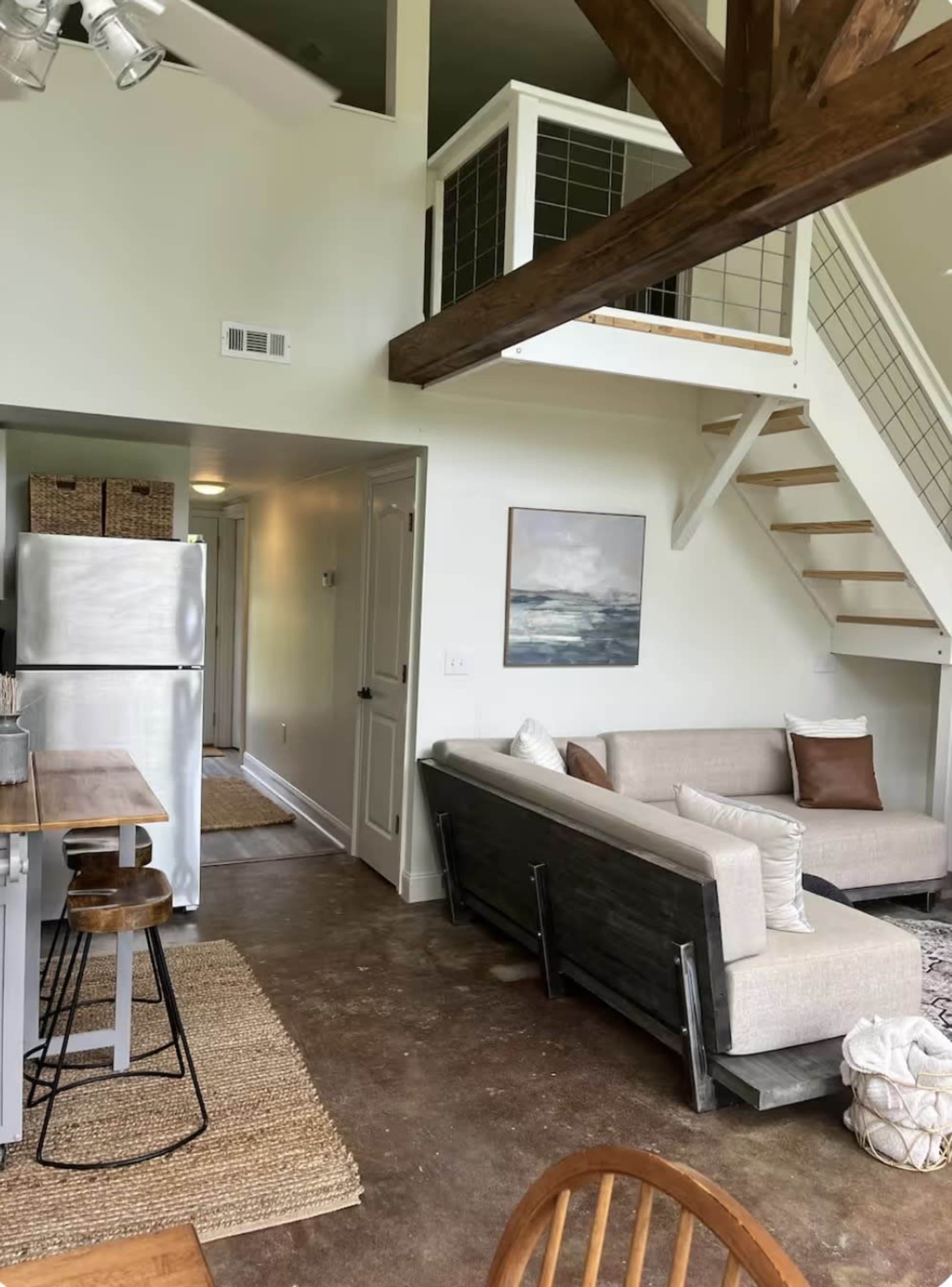 The image shows an open-concept living space featuring a kitchen area with a metal fridge and a dining table, adjacent to a cozy seating area with a sectional sofa and a staircase leading to a loft.