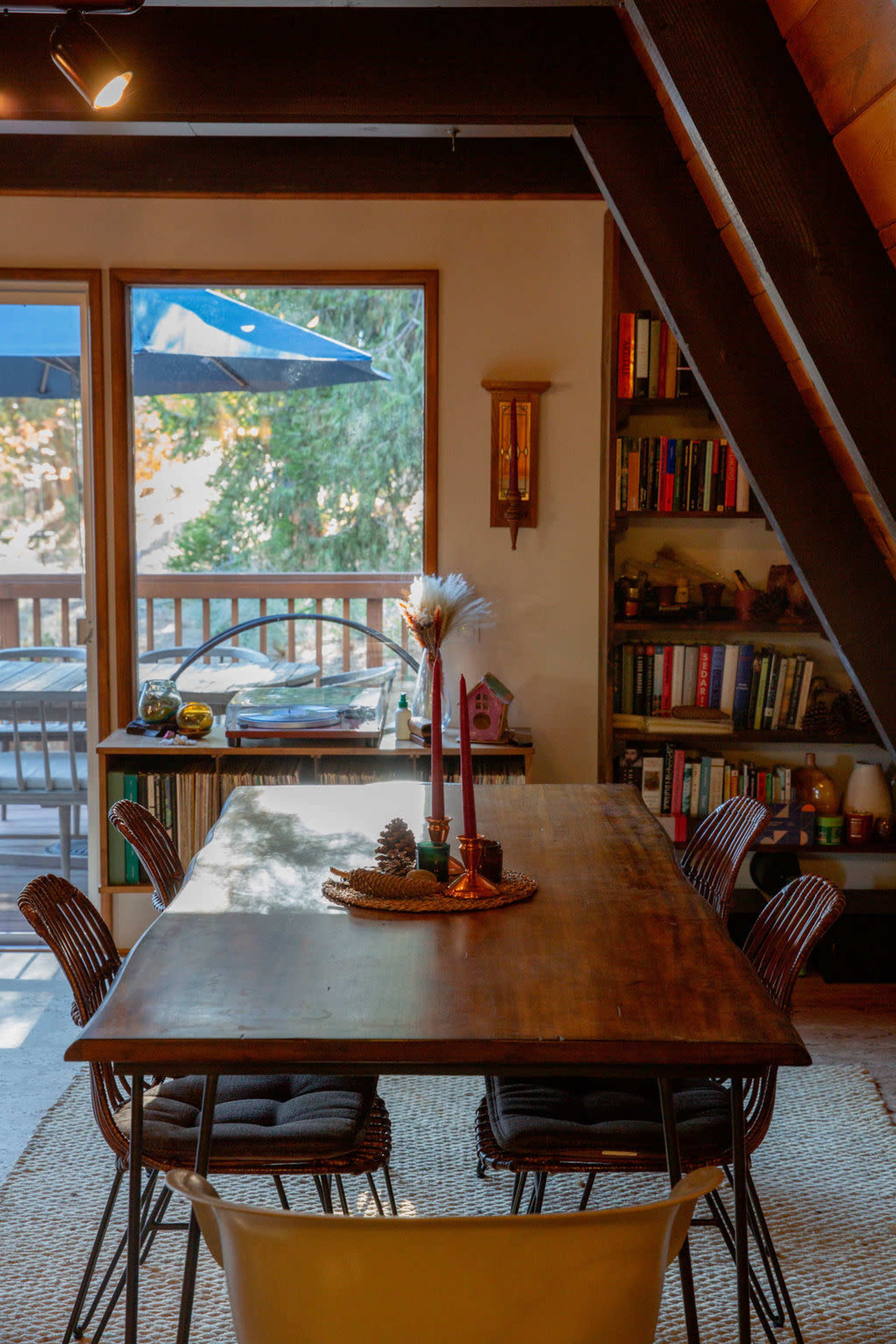 A wooden dining table with candles is set in a well-lit room featuring a bookshelf and large windows that overlook a deck.