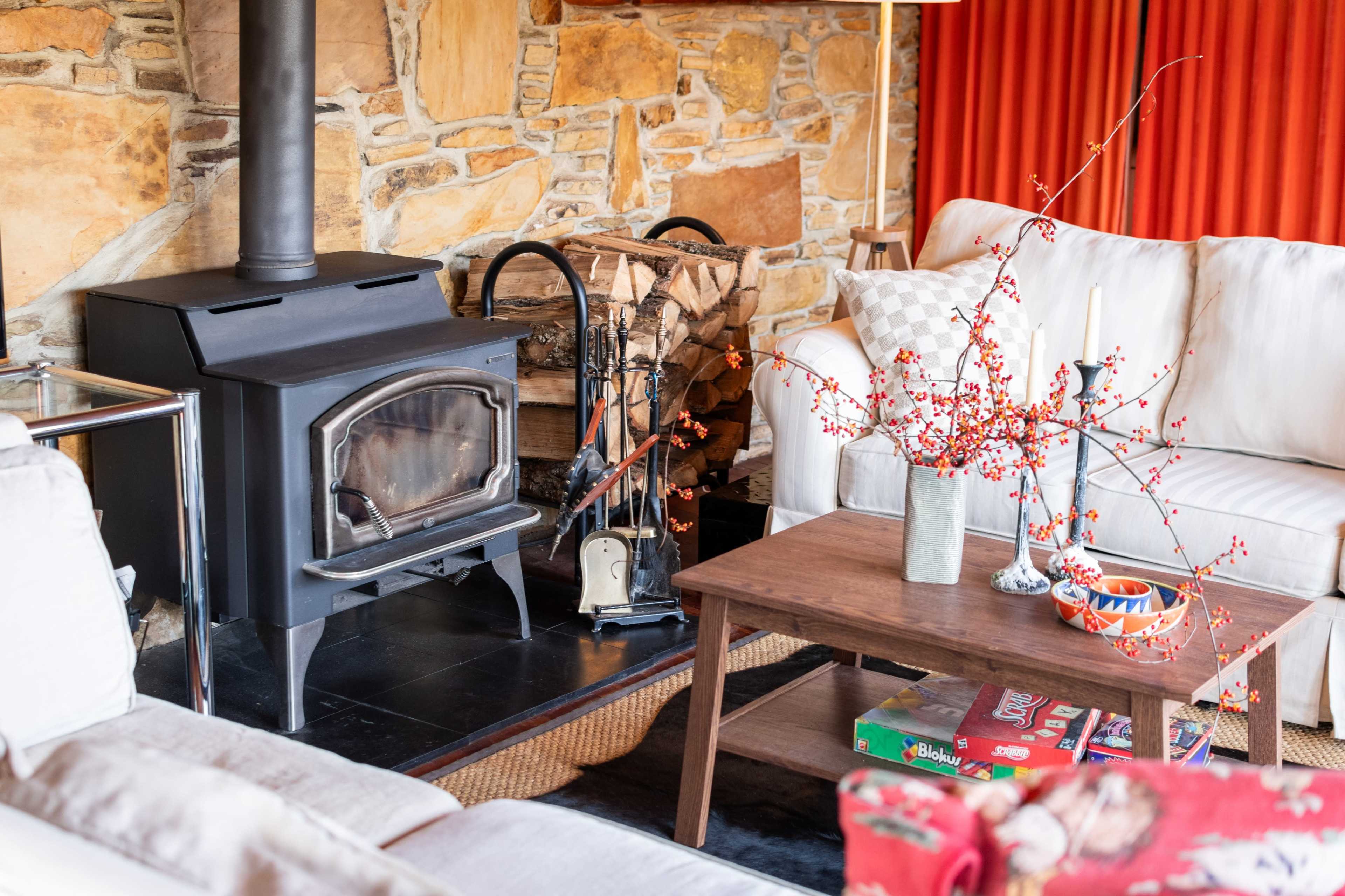 The image shows a cozy living room with a black stove, a stone wall, a wooden coffee table, and a sofa adorned with orange curtains in the background.