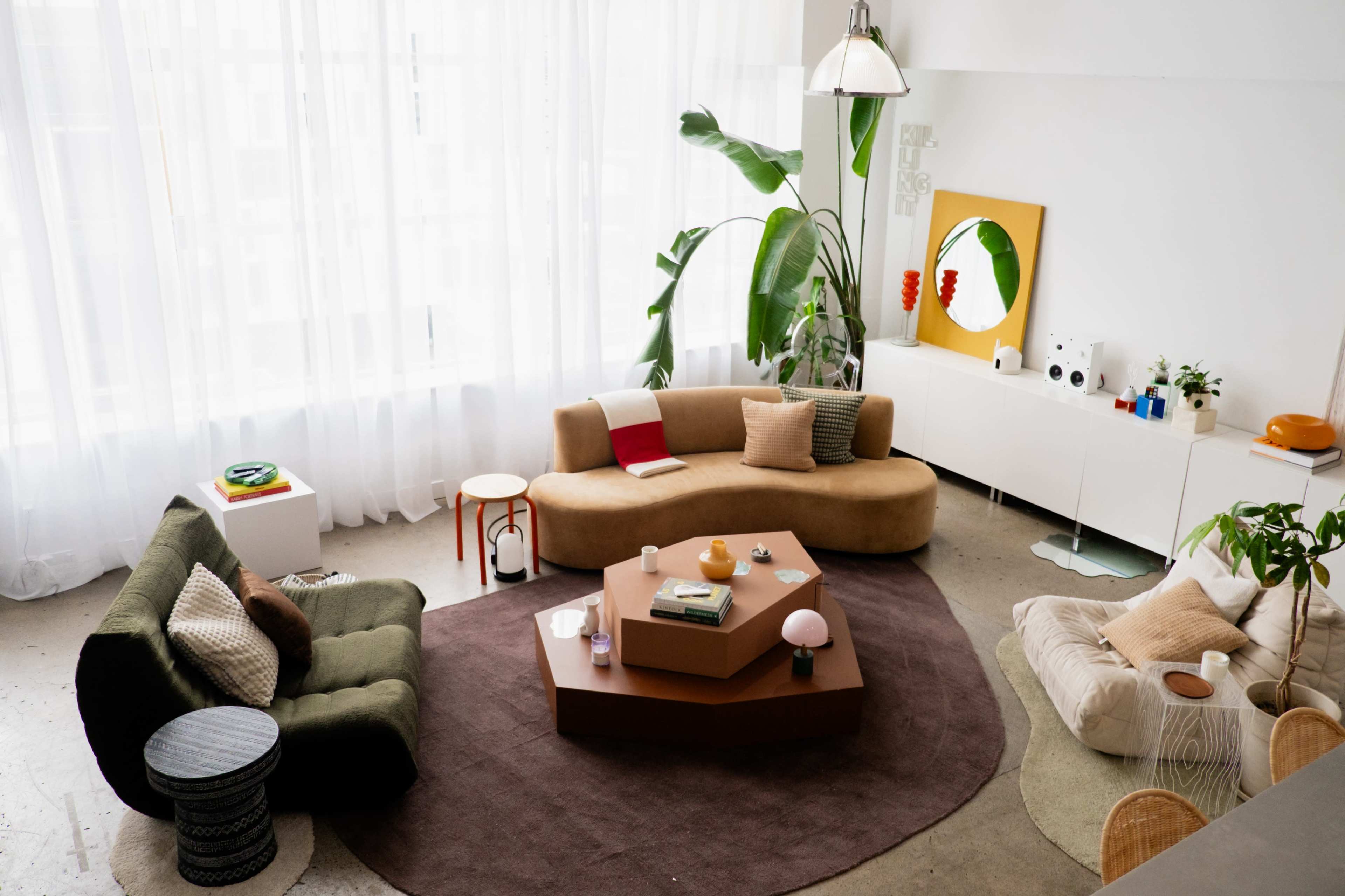 The image shows a modern living room featuring various seating options, a large rug, and bright natural light streaming through sheer curtains.