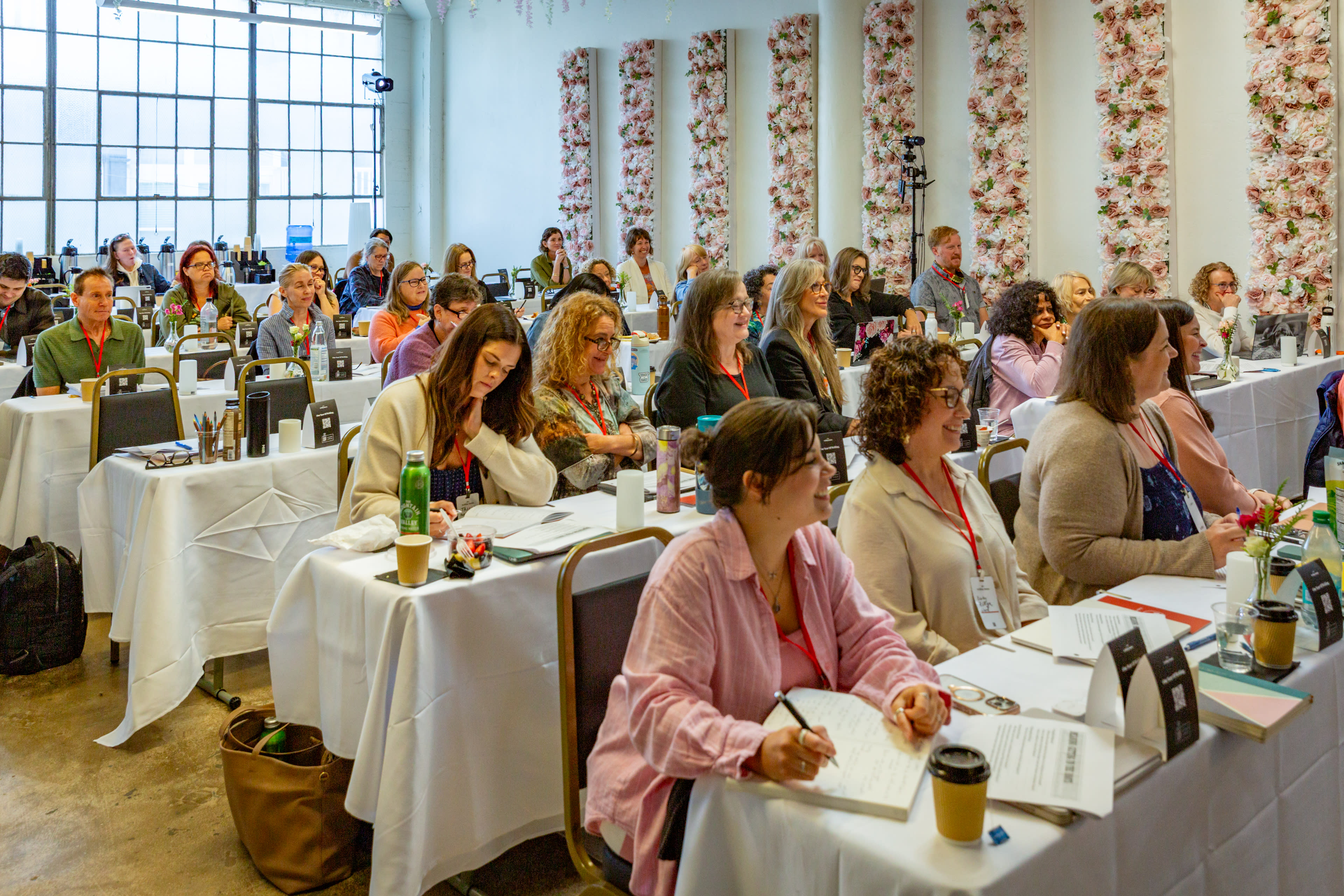 A group of attendees sits at tables in a well-lit conference room, engaged in a presentation, with floral decorations adorning the walls.