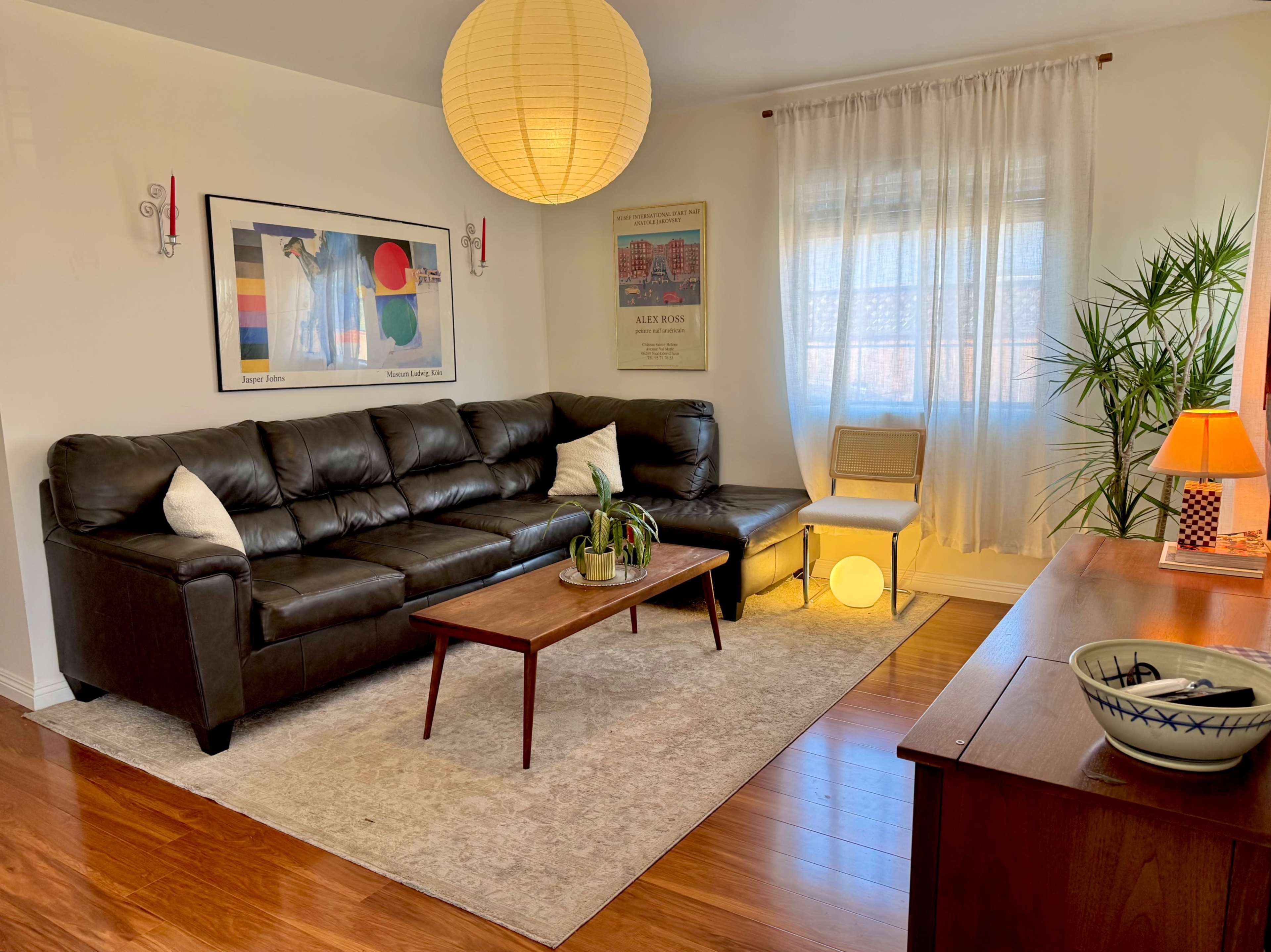 The image shows a well-lit living room featuring a black leather sectional sofa, a wooden coffee table, a rug, and decorative plants.