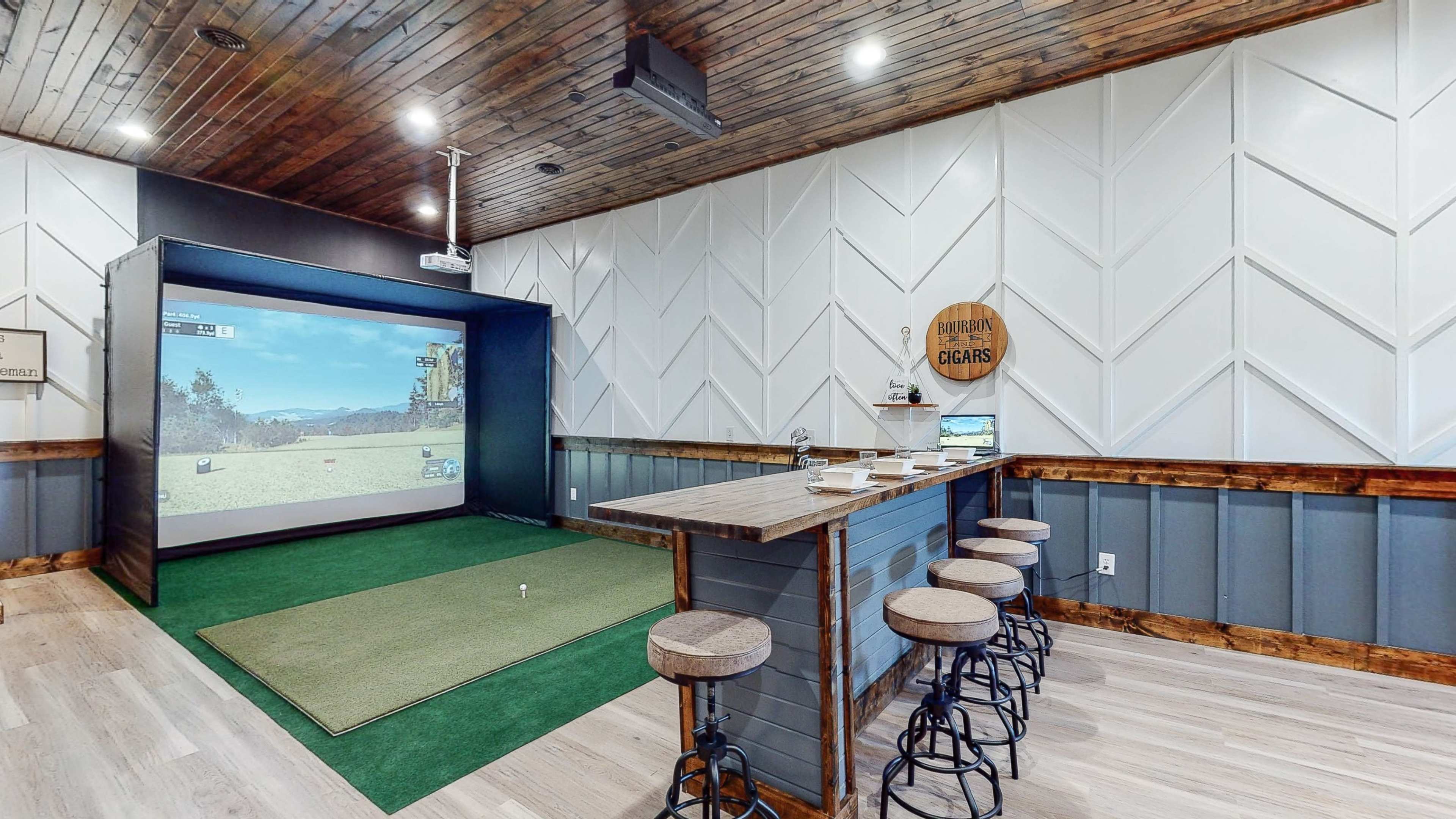 An indoor golf simulation room featuring a large projected screen, a green turf area, and a bar with stools.