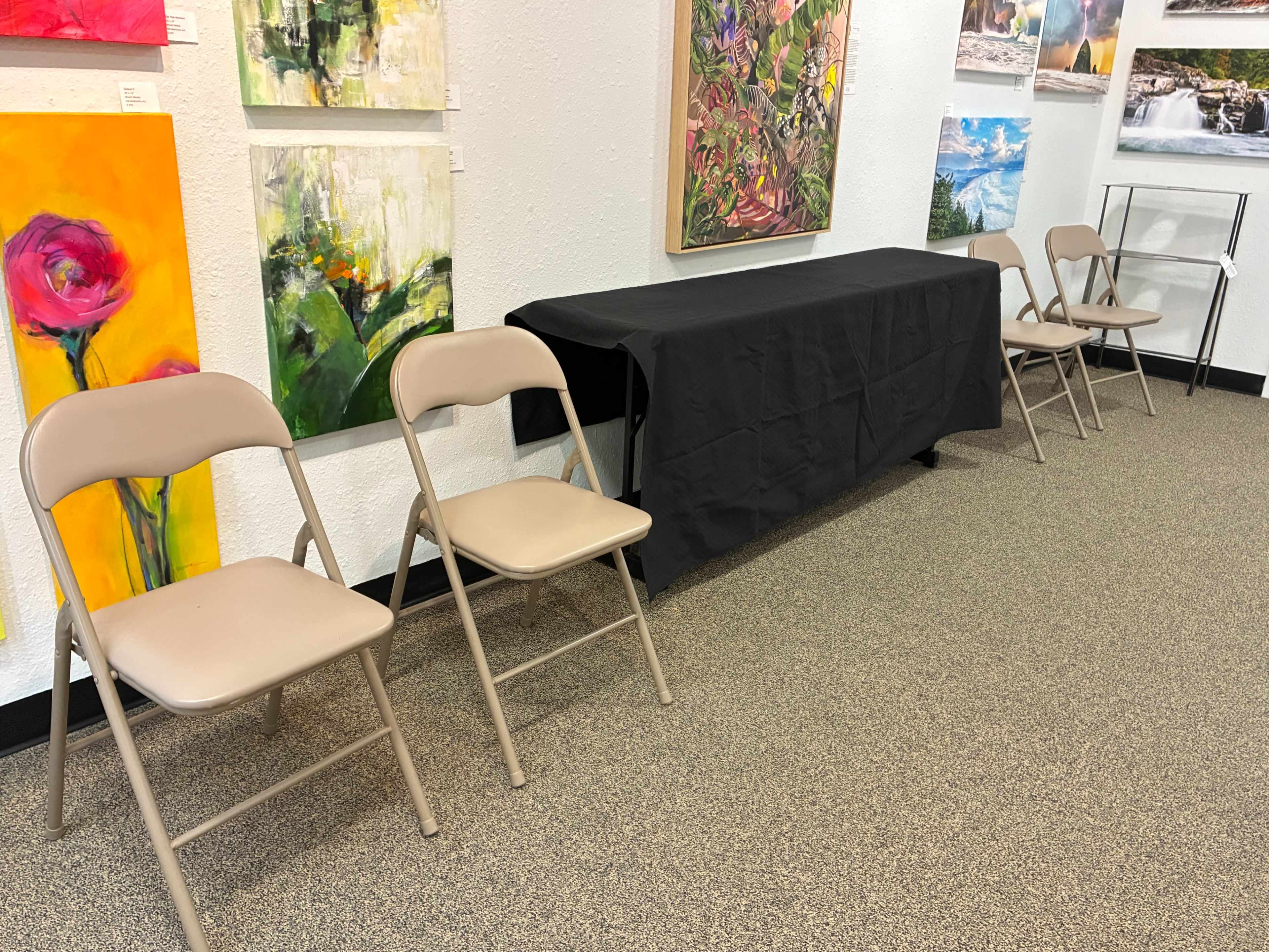 The image shows a small exhibition space with a table covered in a black cloth and three beige folding chairs arranged in front of it, against a backdrop of colorful artwork on the walls.