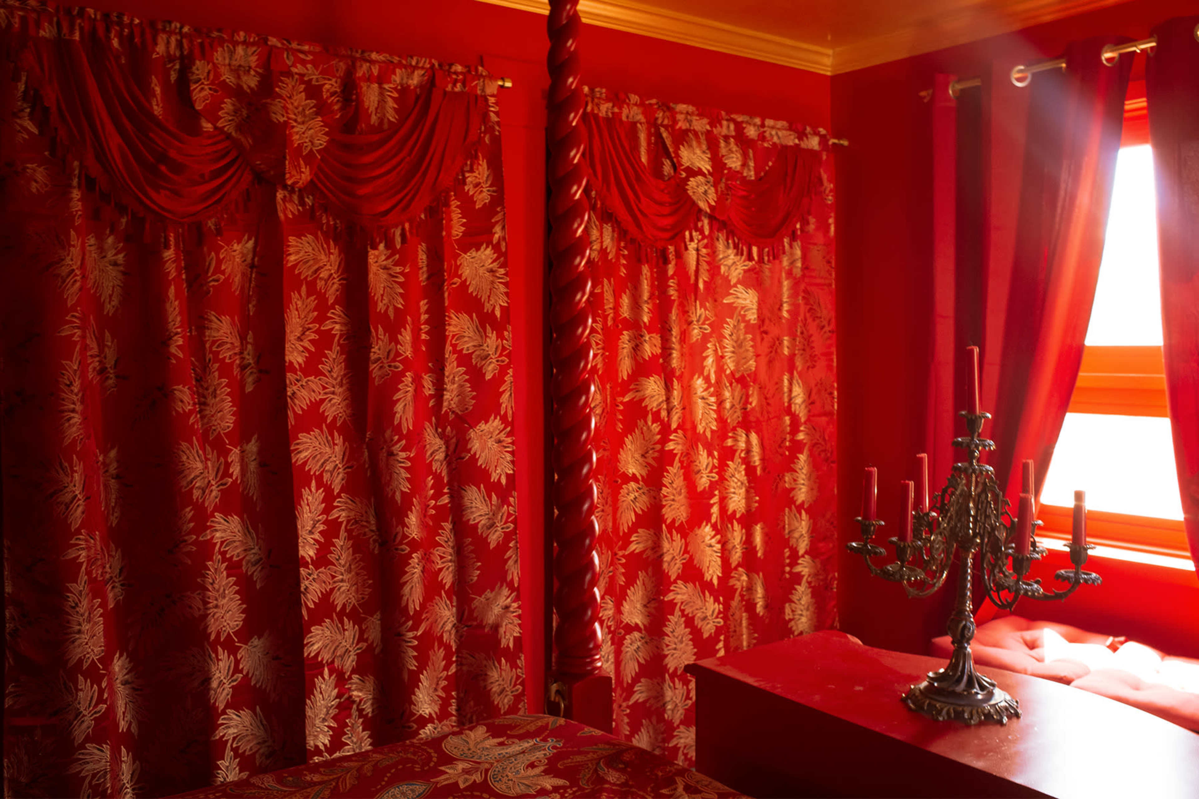 The scene features a bedroom with red curtains, a red bedspread, and a decorative candelabra on a red dresser.