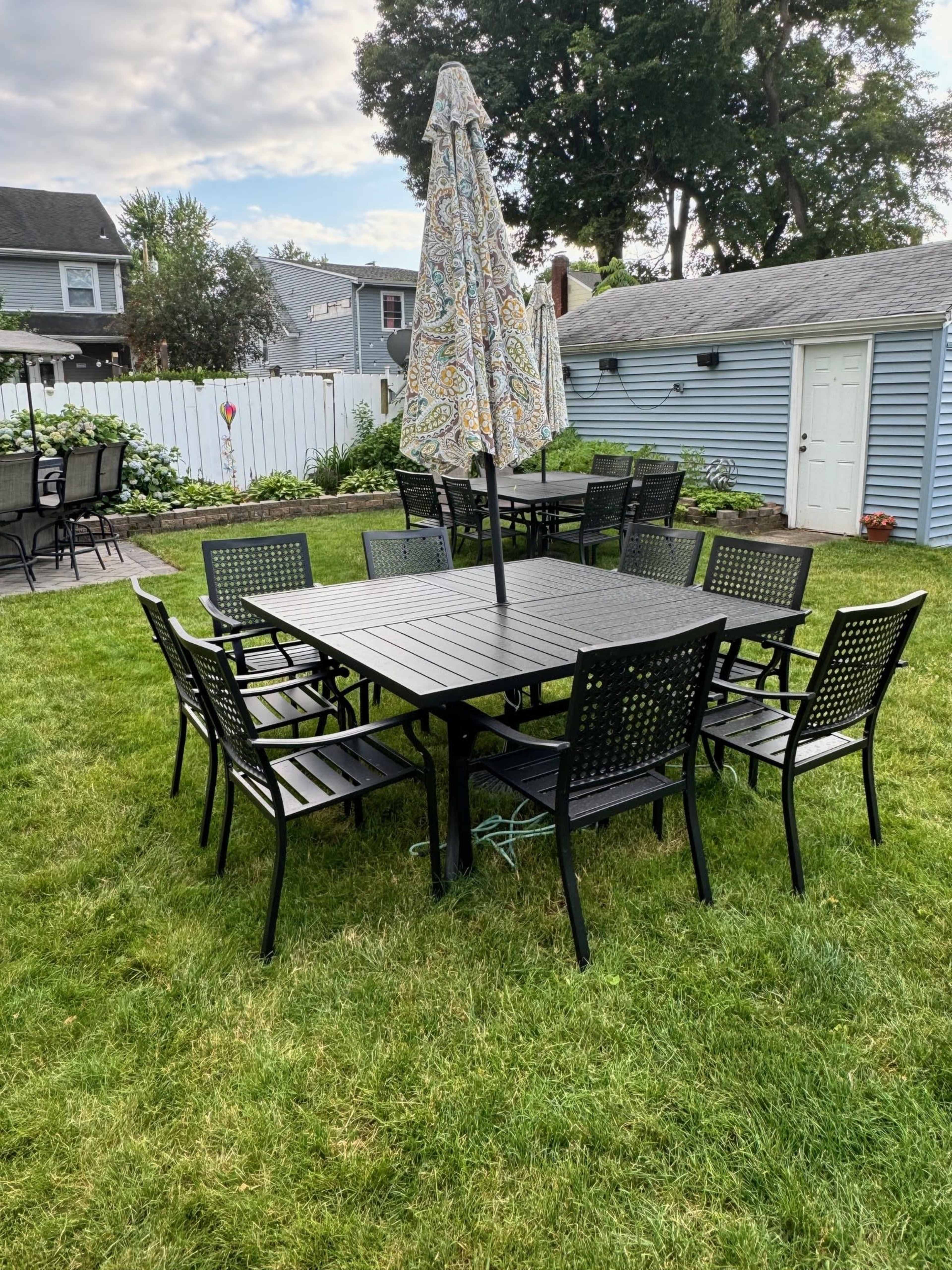 A rectangular outdoor dining table with eight chairs is set on a grassy area, shaded by a large umbrella.