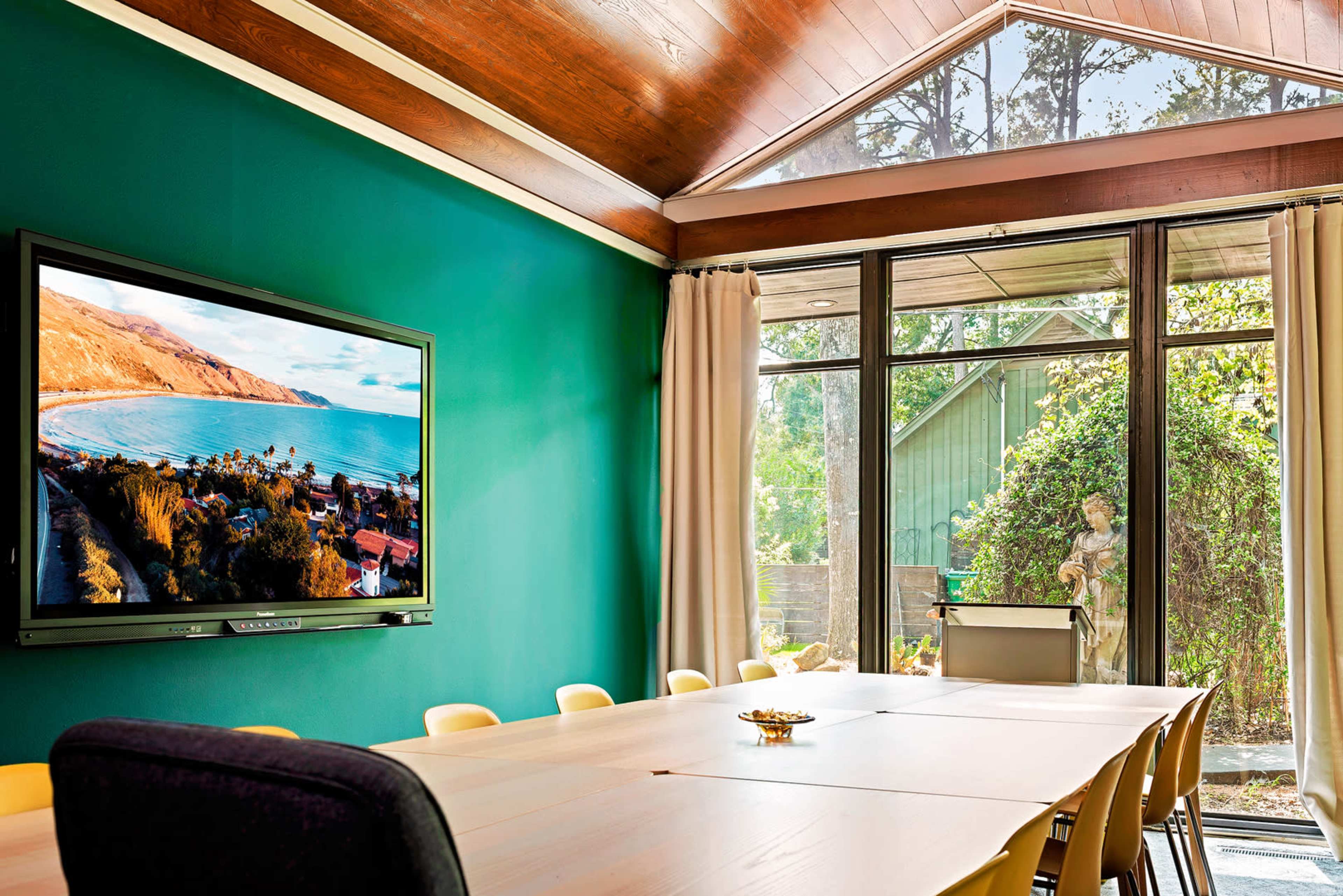 The image shows a conference room with a large table, a television mounted on the wall displaying a coastal view, and large windows allowing natural light to enter the space.