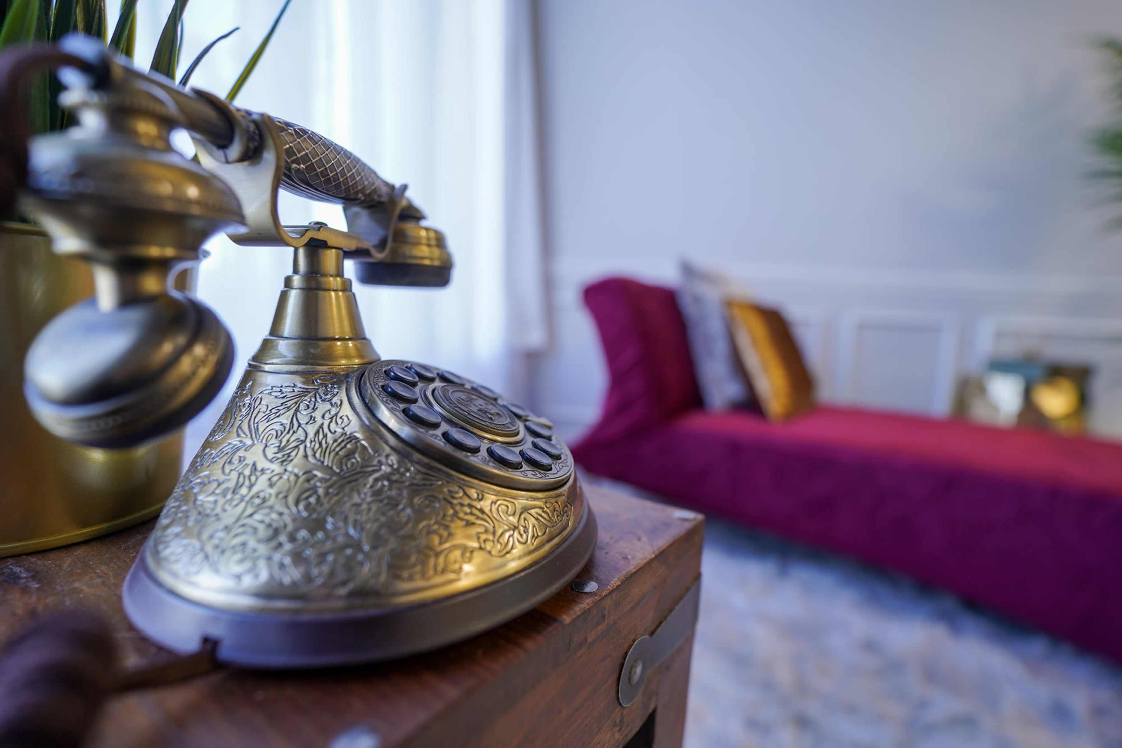 An ornate vintage telephone is placed on a wooden table, with a plush red chaise lounge and decorative pillows in the background.