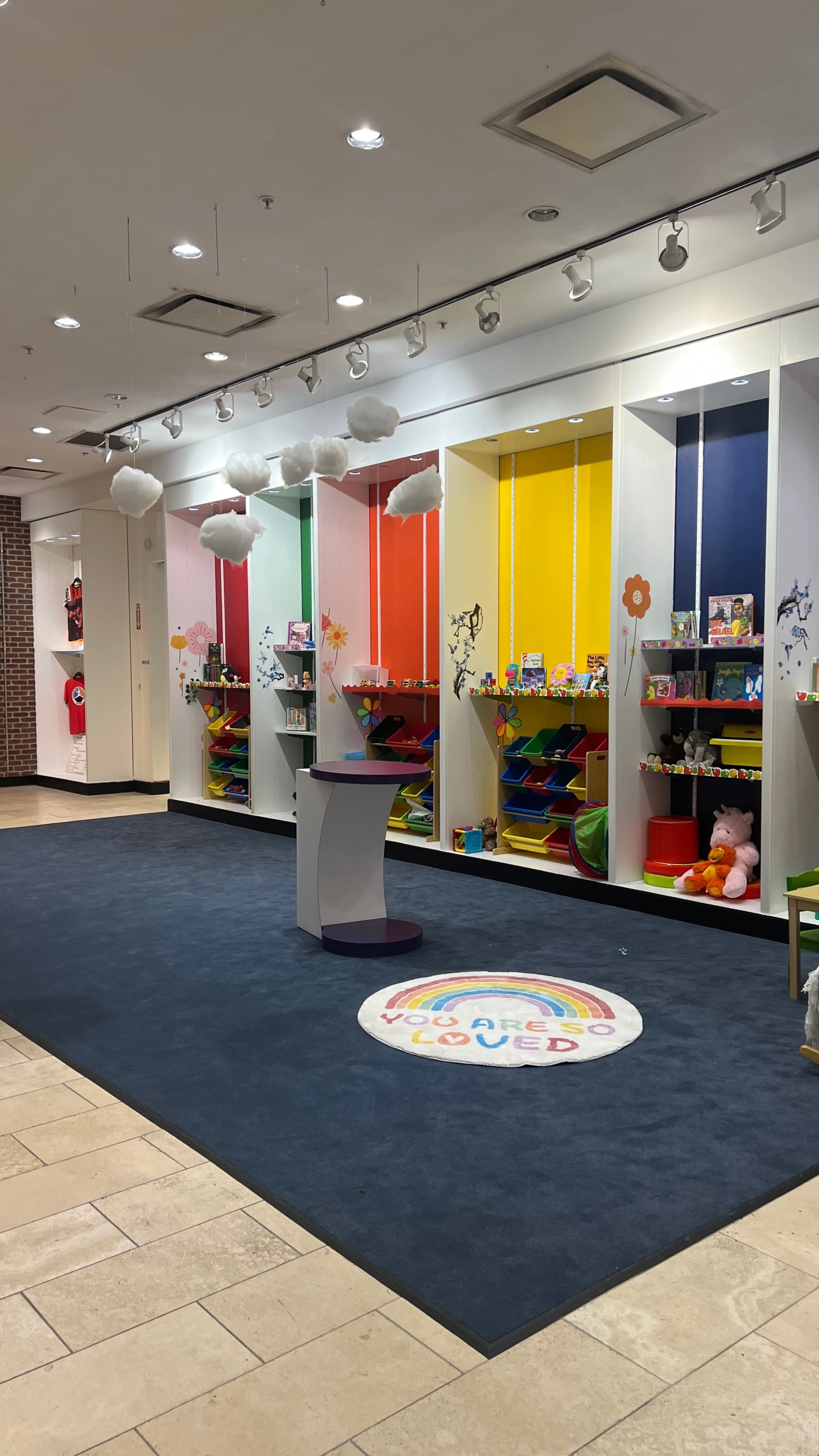The image shows a brightly colored children's play area with several shelving units filled with toys and a circular rug on the floor featuring a rainbow design.