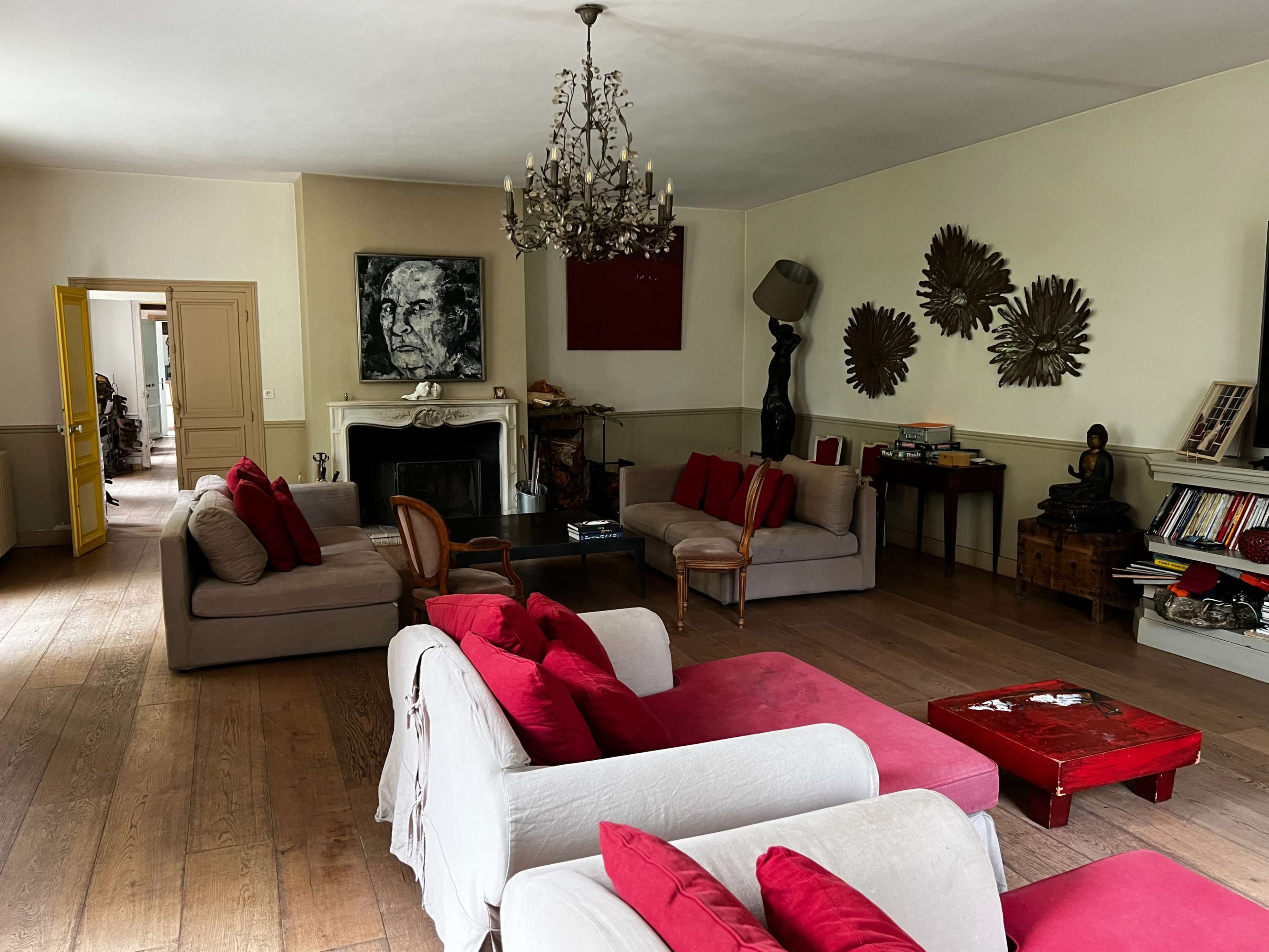 A spacious living room furnished with beige sofas adorned with red cushions, a chandelier hanging from the ceiling, and various decorative items on the walls.