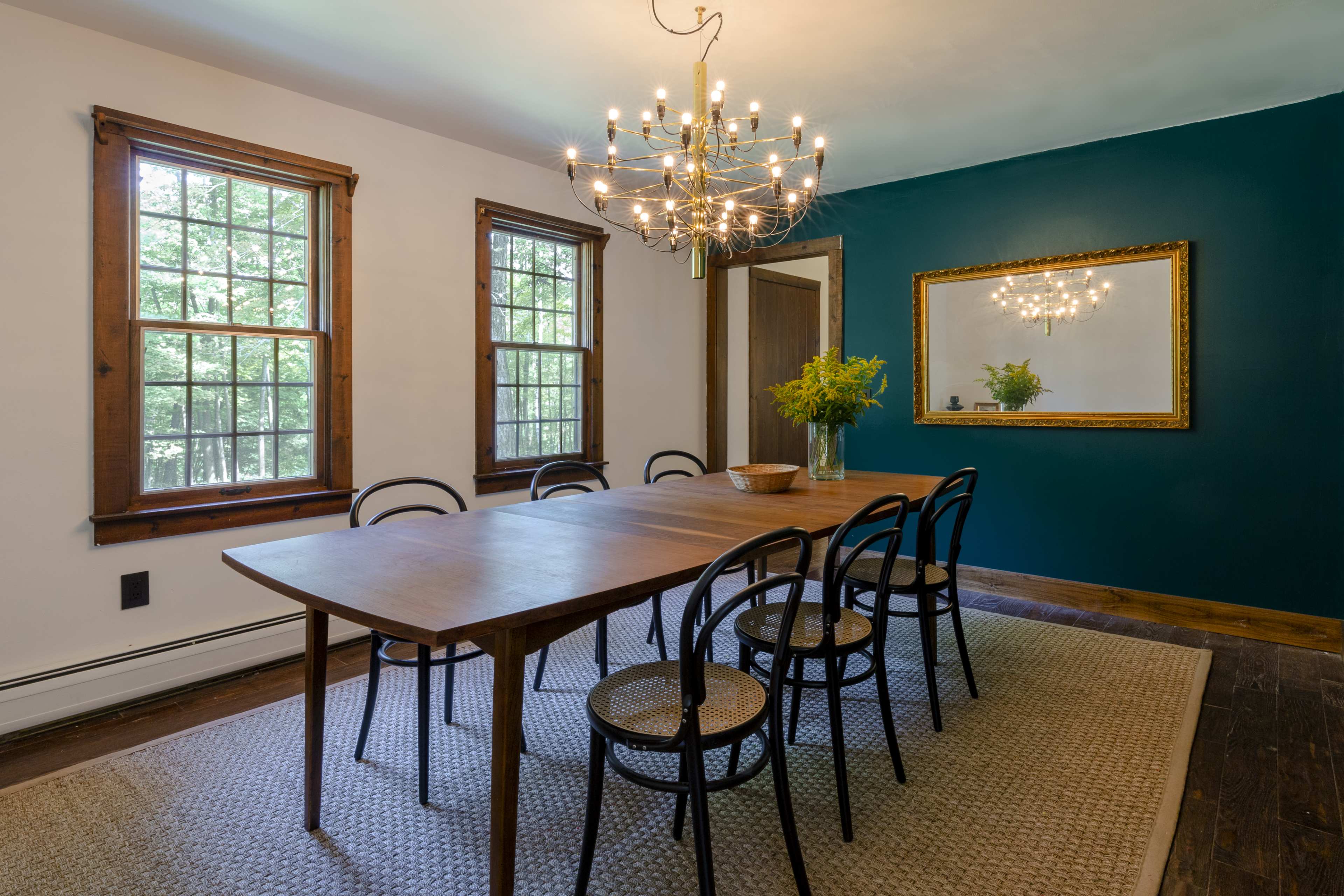 A dining room featuring a long wooden table surrounded by six chairs, illuminated by a chandelier, with large windows letting in natural light and a dark green accent wall.