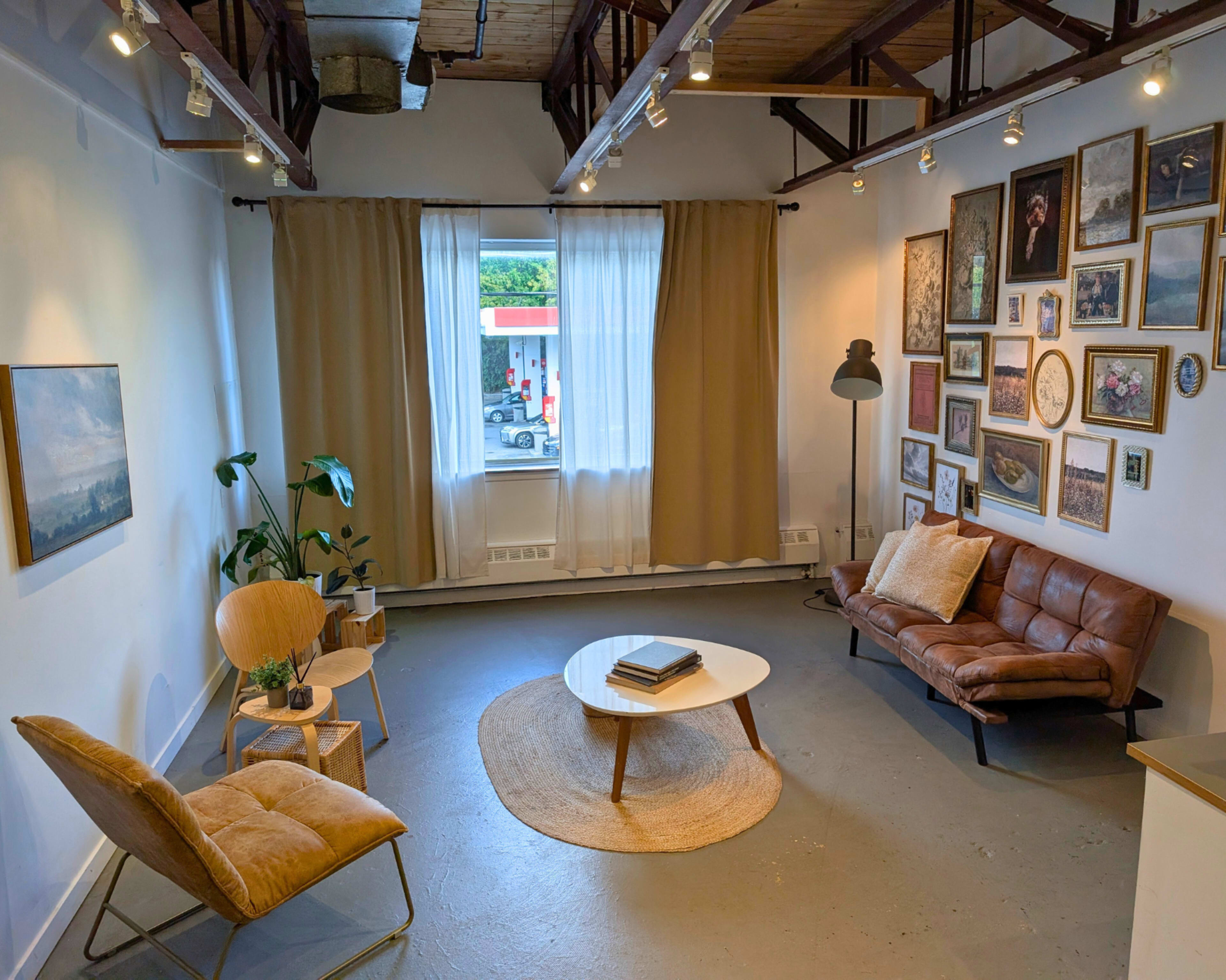 A cozy room features a brown sofa and two chairs encircling a white coffee table, with decorative art pieces on the walls and natural light streaming in through large windows.
