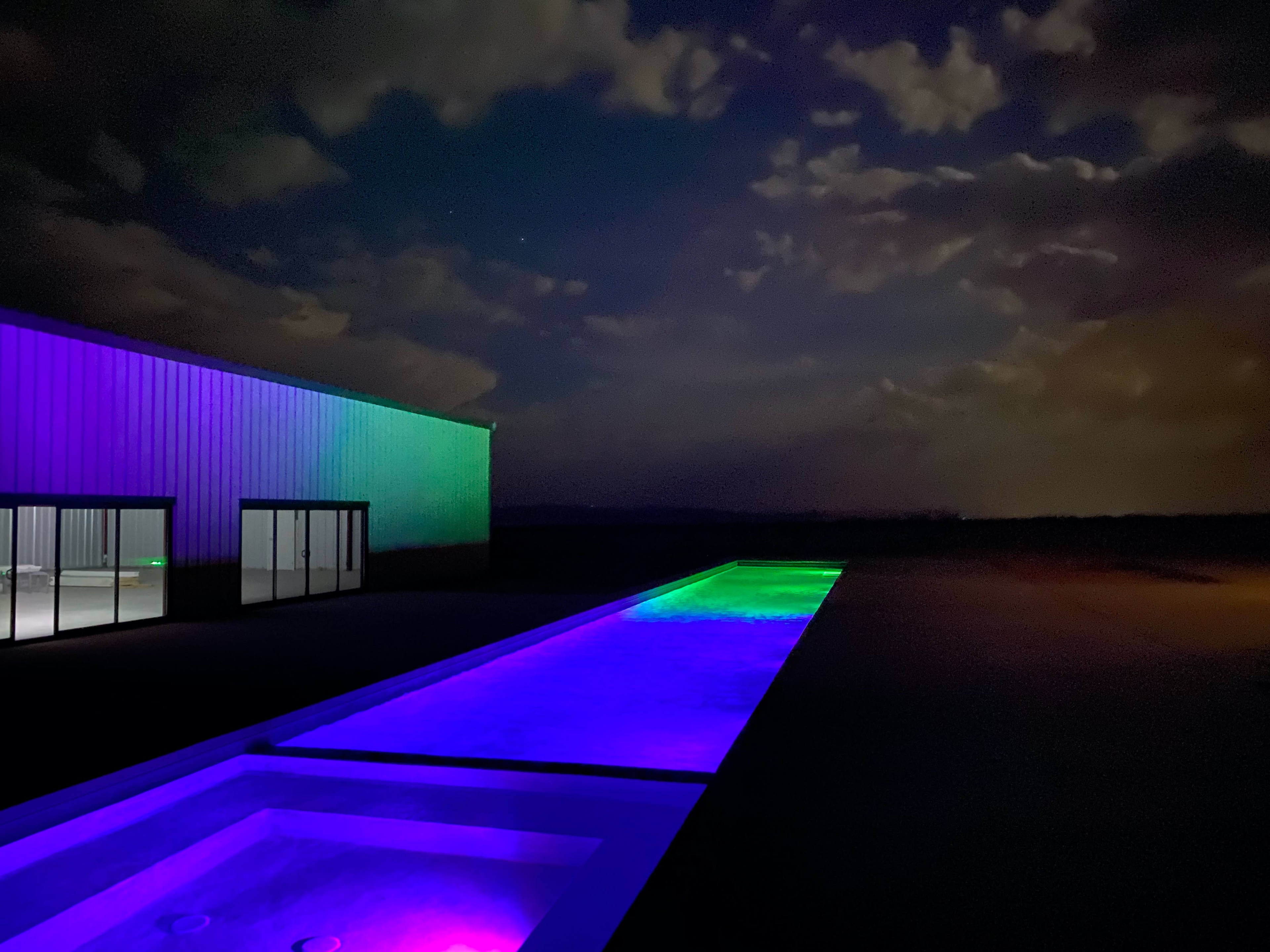 A brightly lit pool area with colored lights runs alongside a large building under a night sky.