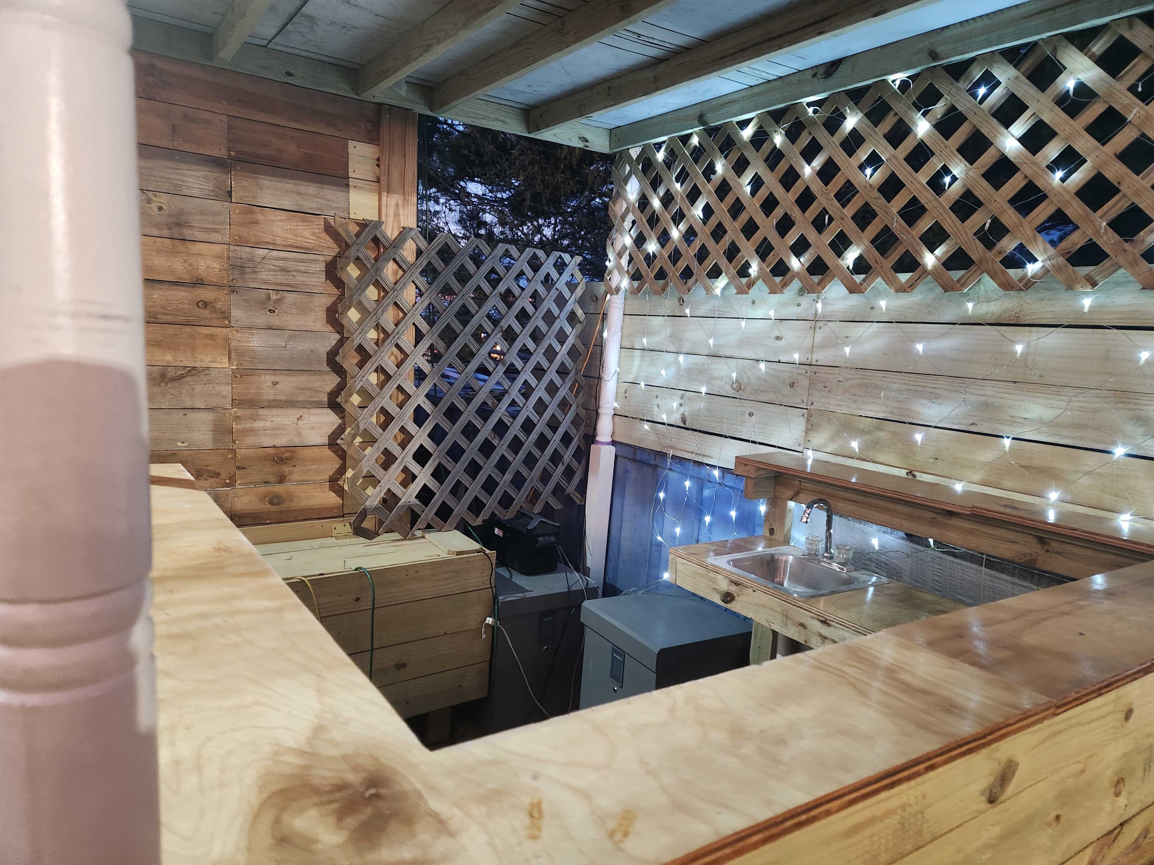 A wooden outdoor space featuring a bar area with a lattice wall, a sink, and string lights illuminating the surroundings.