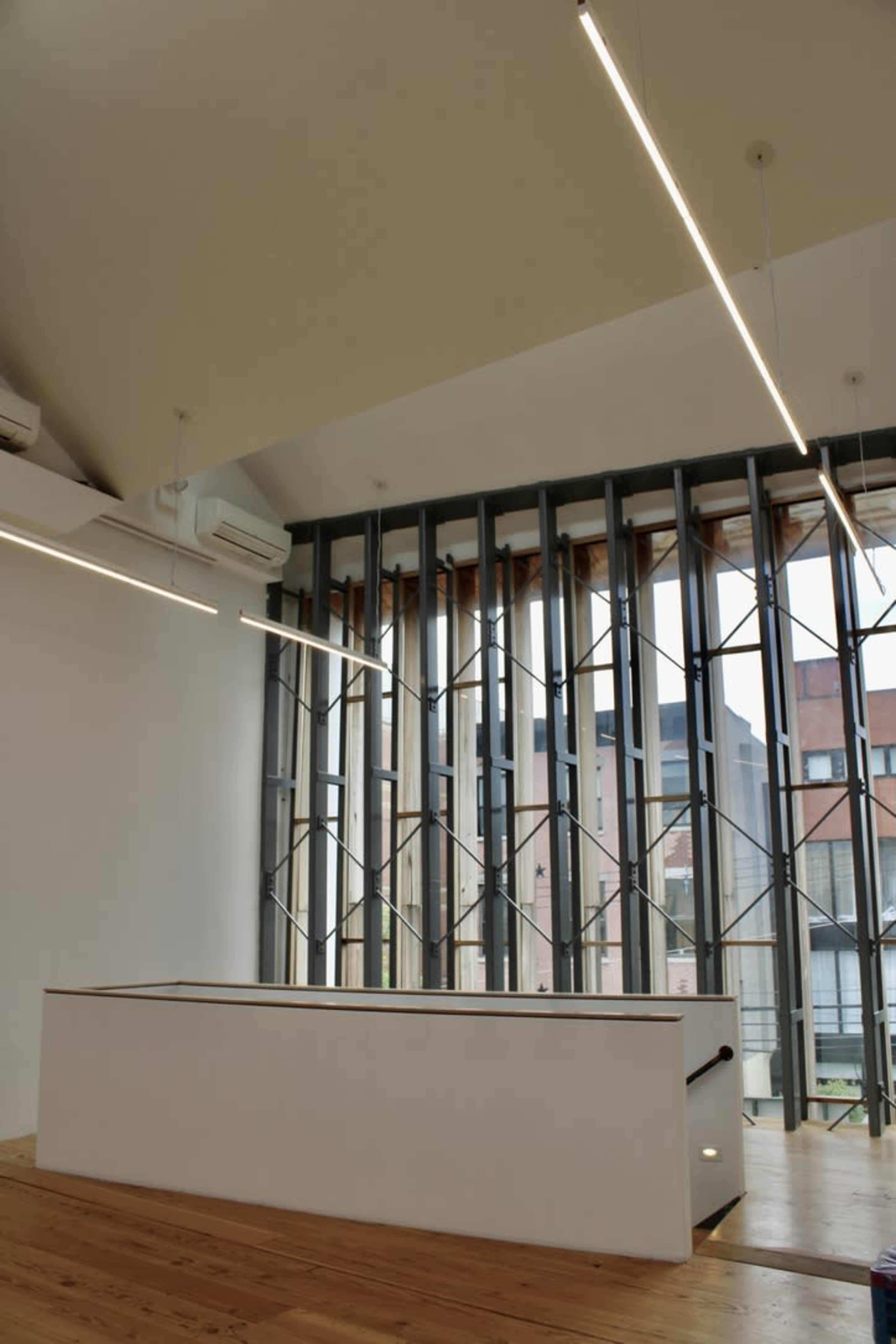 The image shows a modern interior space with an angular ceiling, a white reception desk, and large windows featuring a geometric metal design.