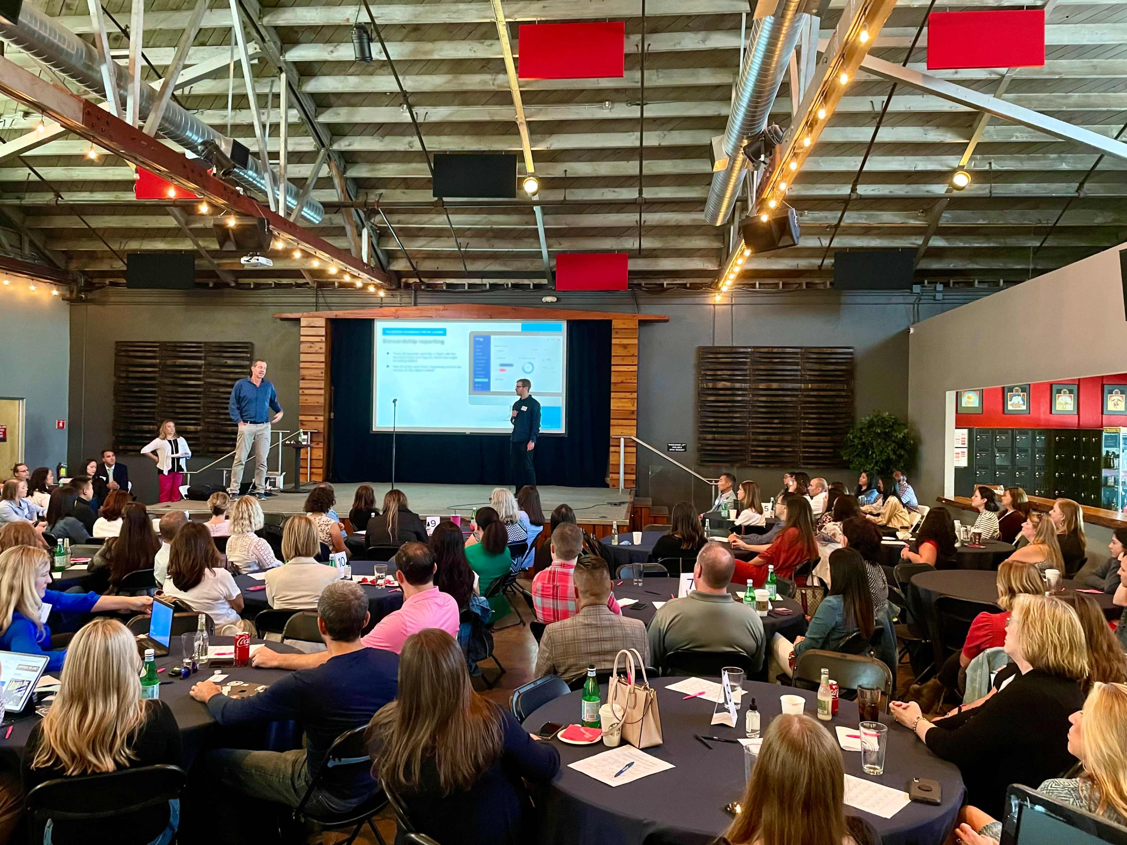 A group of people is seated at tables in a large room, attentively watching a presentation on a screen while two speakers stand at the front.