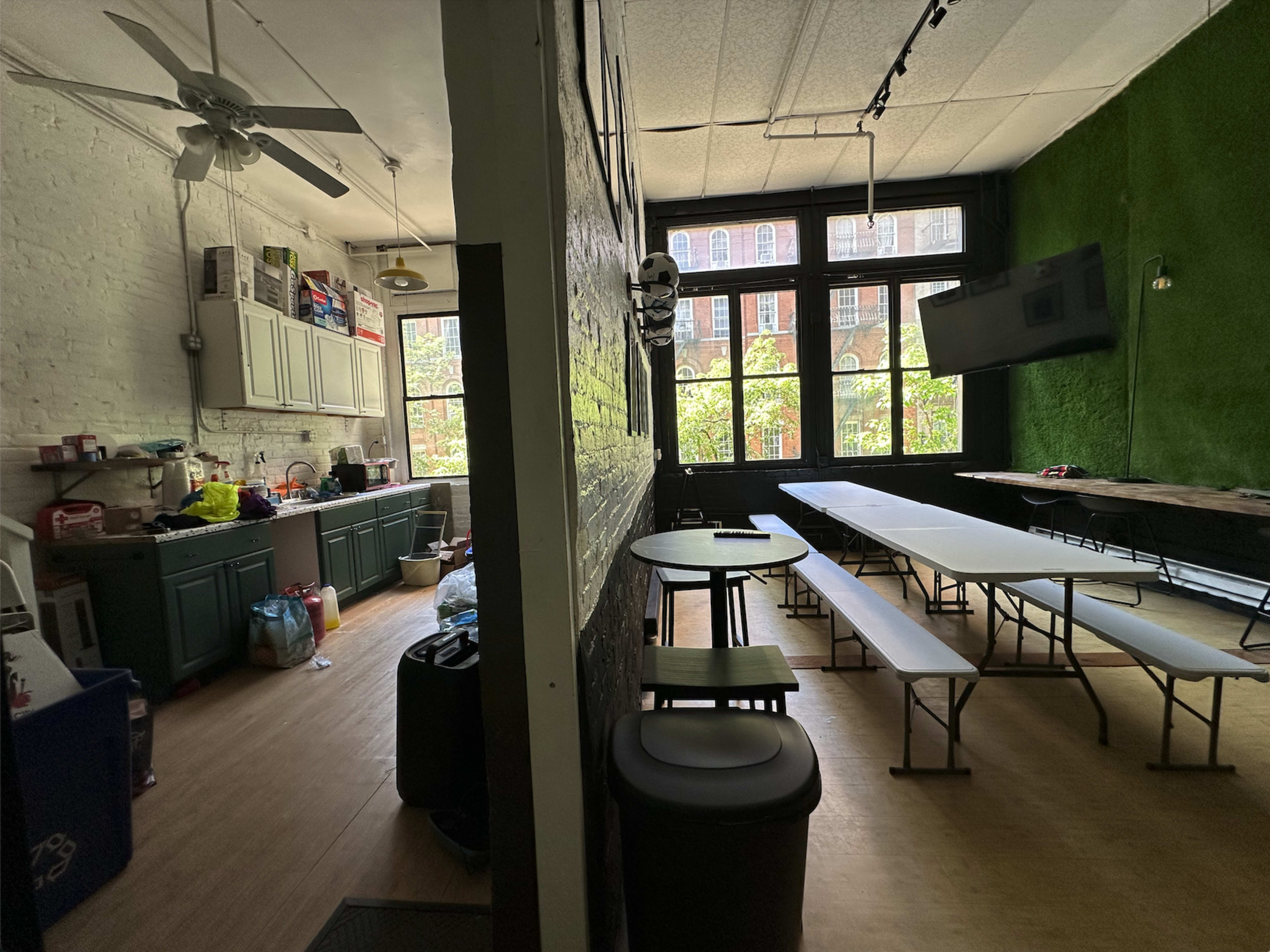 The image shows a room divided into two areas, one featuring a kitchen with cabinets and a sink, and the other containing several tables and chairs arranged for dining or meetings.