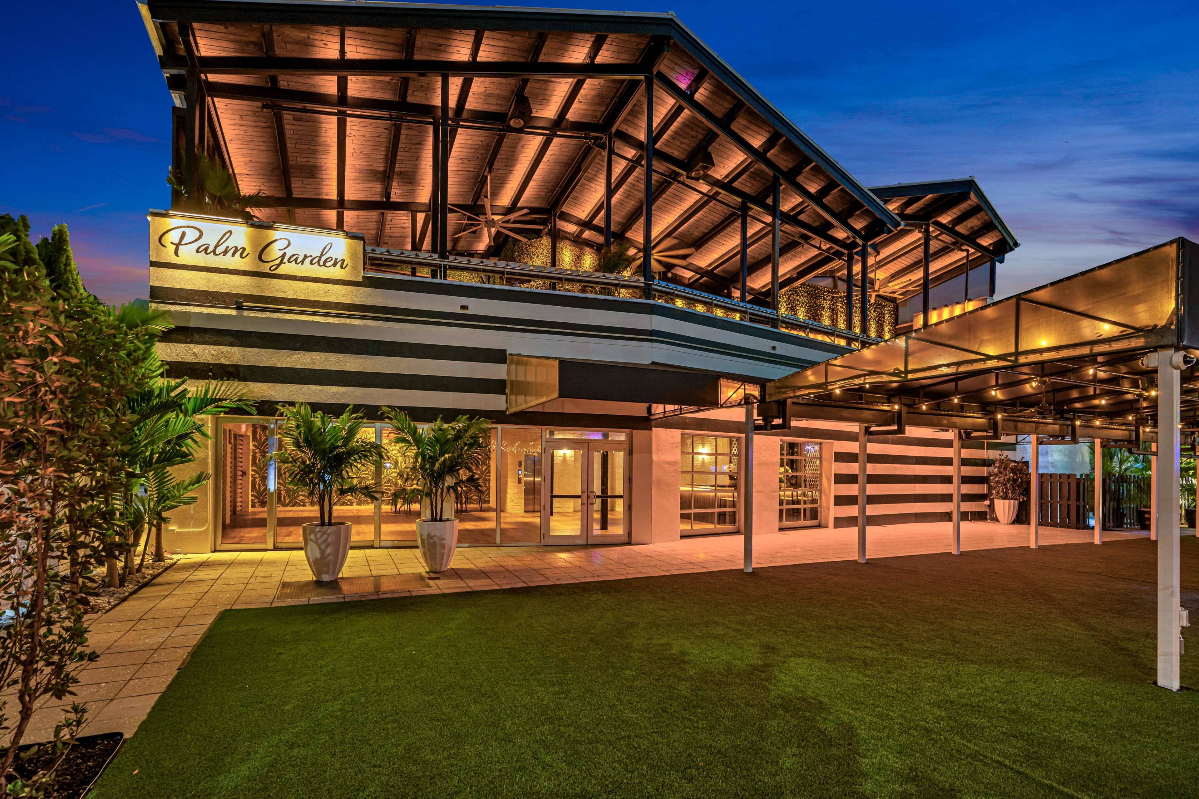 The image shows a well-lit outdoor area of a building named "Palm Garden," featuring a wooden structure with a spacious patio and landscaped greenery.