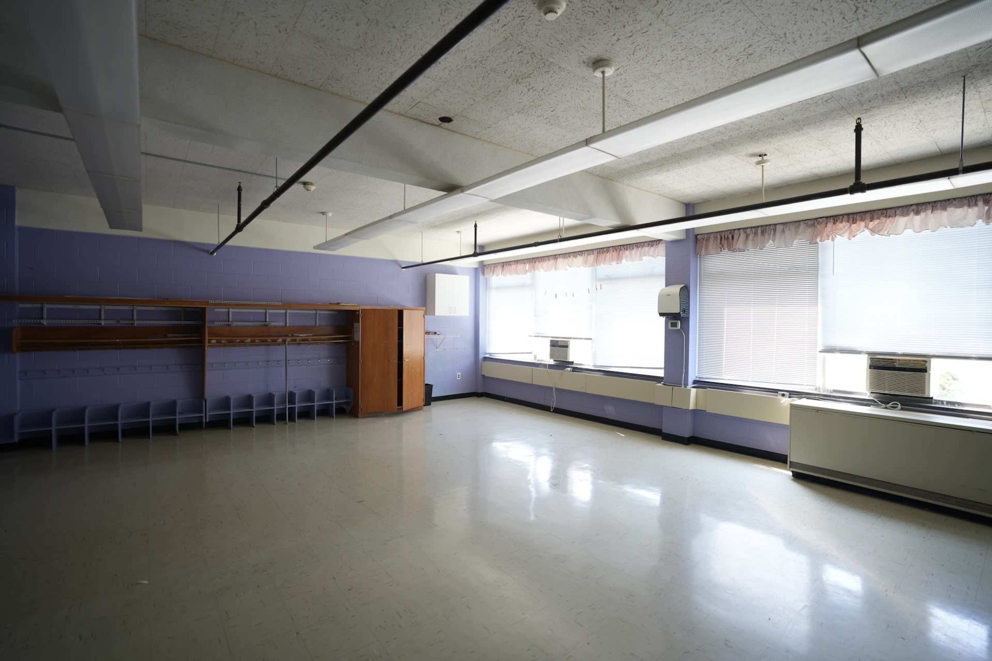 The image shows an empty classroom with purple walls, large windows, and a row of hooks along one side.