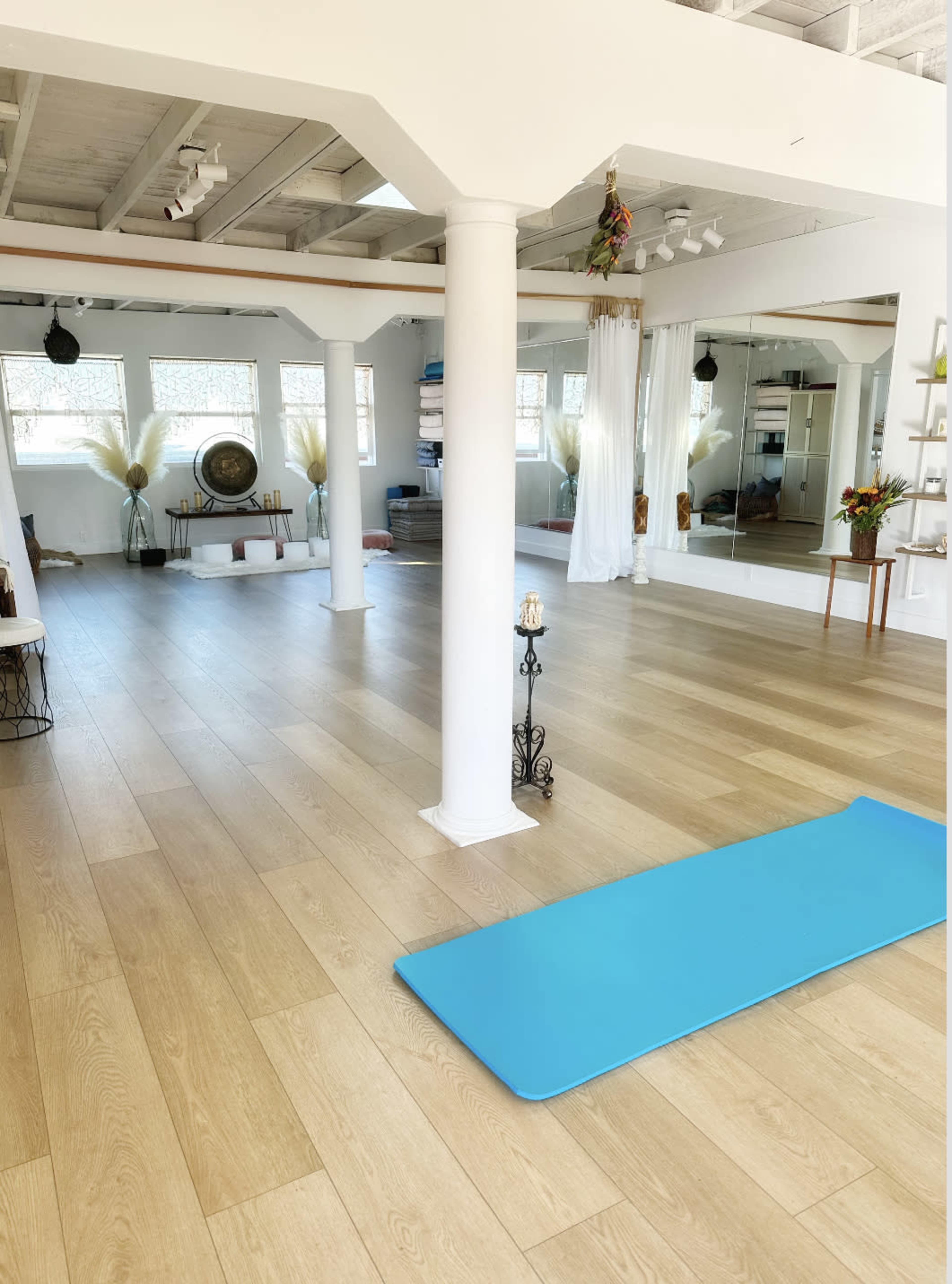 The image shows a spacious, well-lit yoga studio with light wood flooring, a blue yoga mat, and large mirrors along one wall.