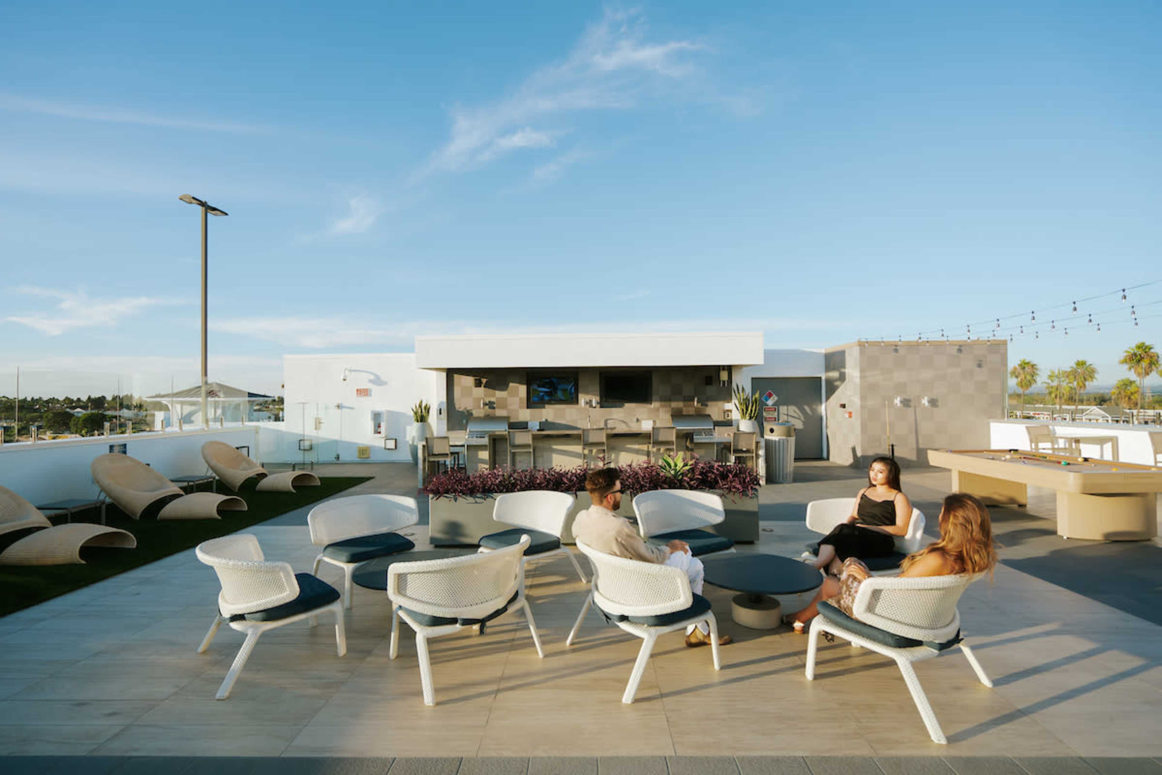 A group of people sits around a circular table on a rooftop patio with a bar and lounge chairs in the background.