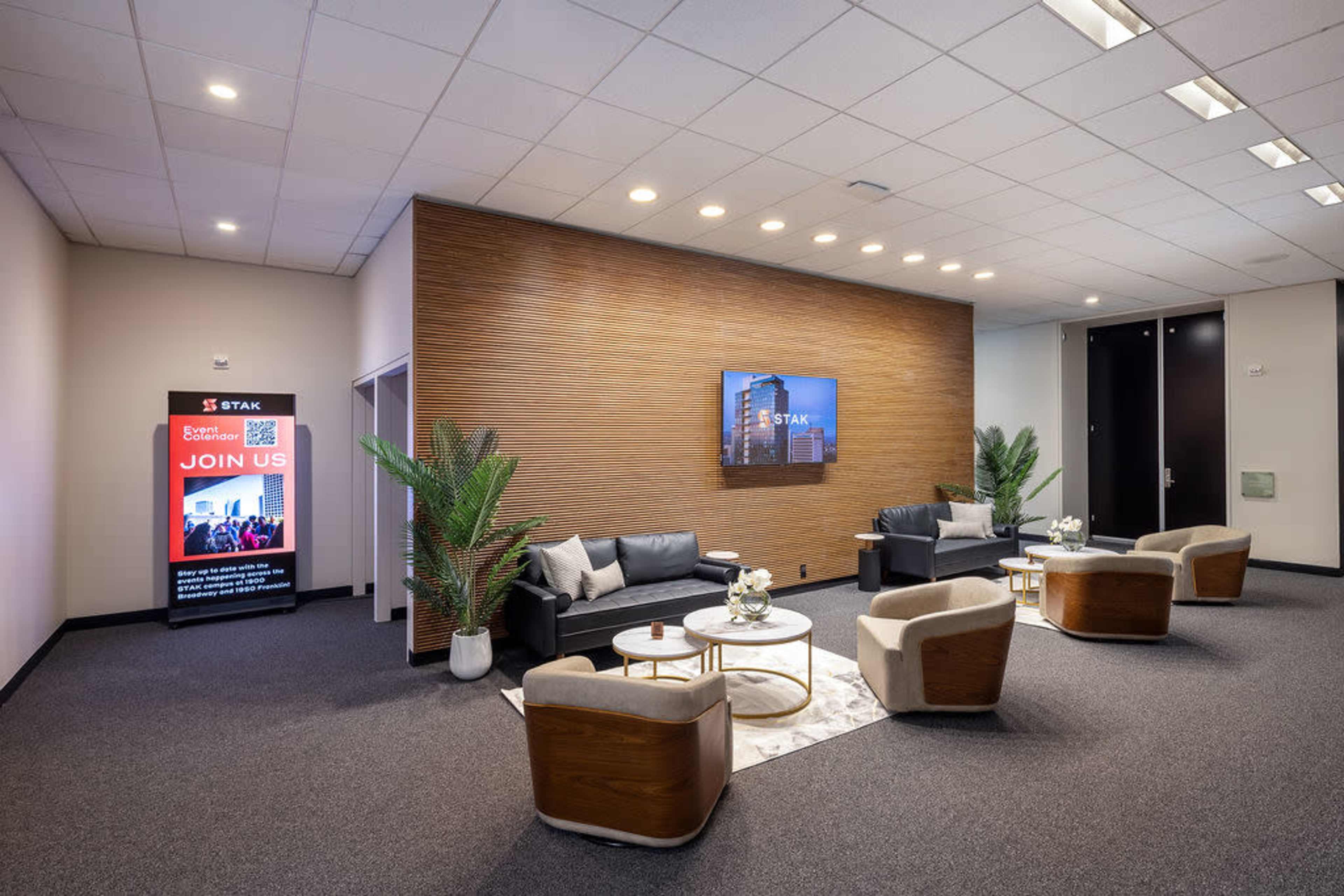 The image shows a modern waiting area with black leather sofas, light-colored armchairs, a marble coffee table, a decorative plant, and a digital advertisement for a business on the wall.