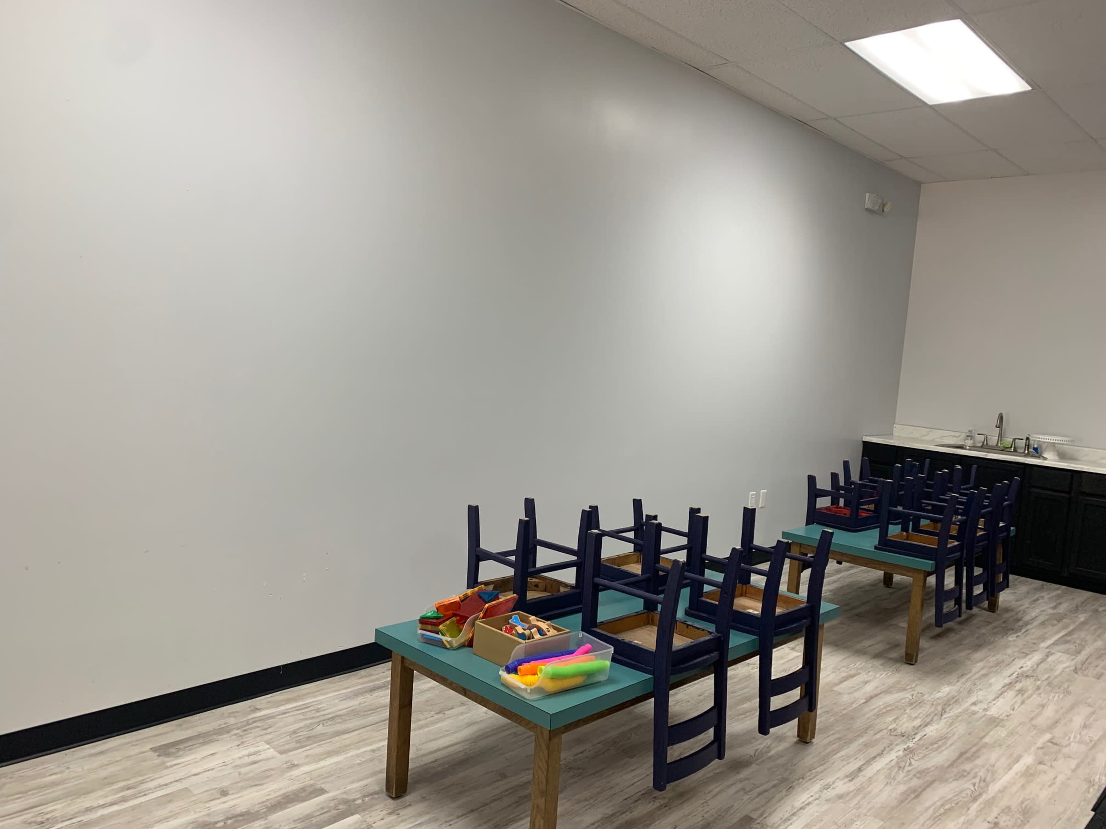 An empty classroom features two tables with chairs stacked on top, and a kitchenette area in the background.