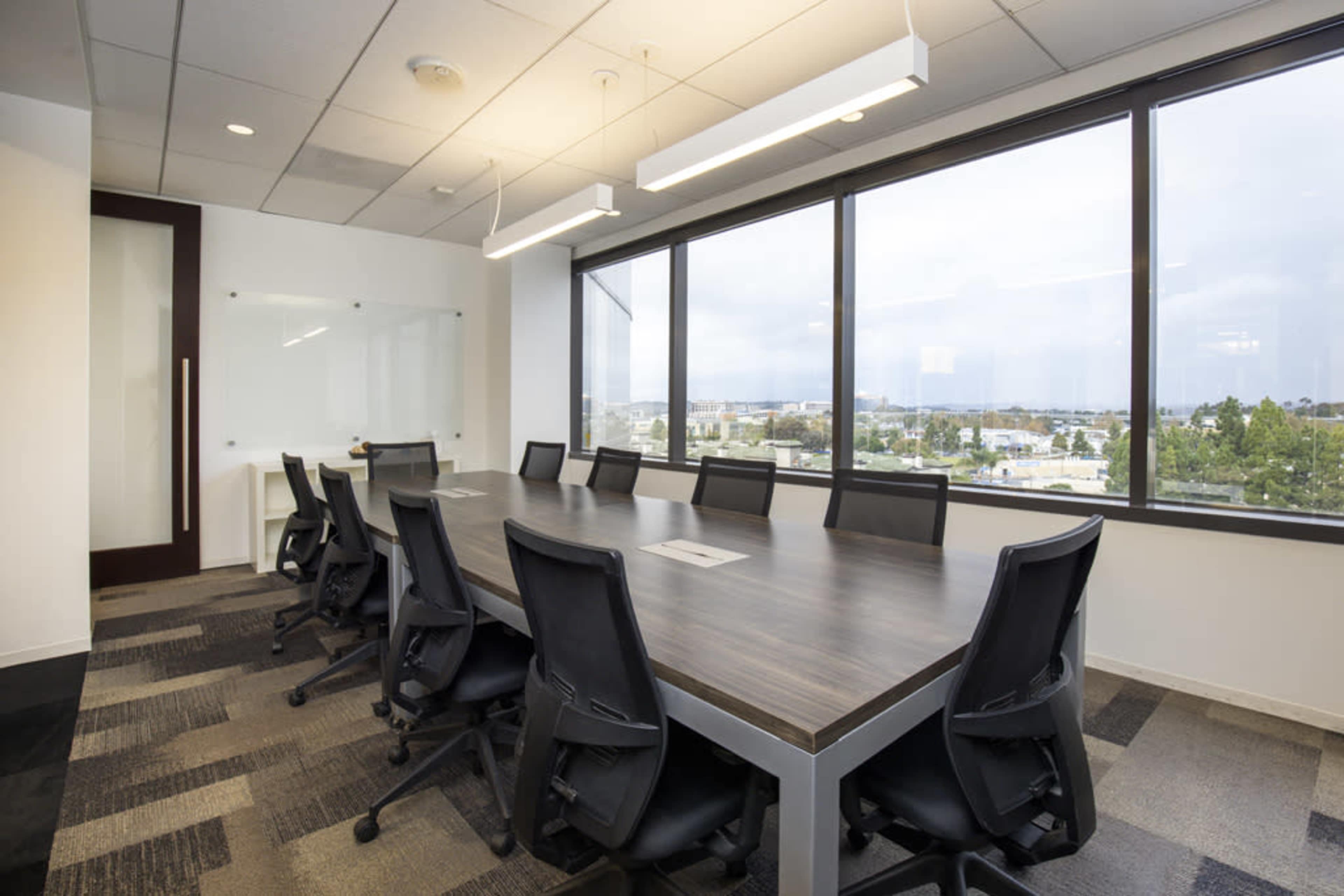 A conference room features a long wooden table surrounded by black chairs and large windows overlooking a cityscape.