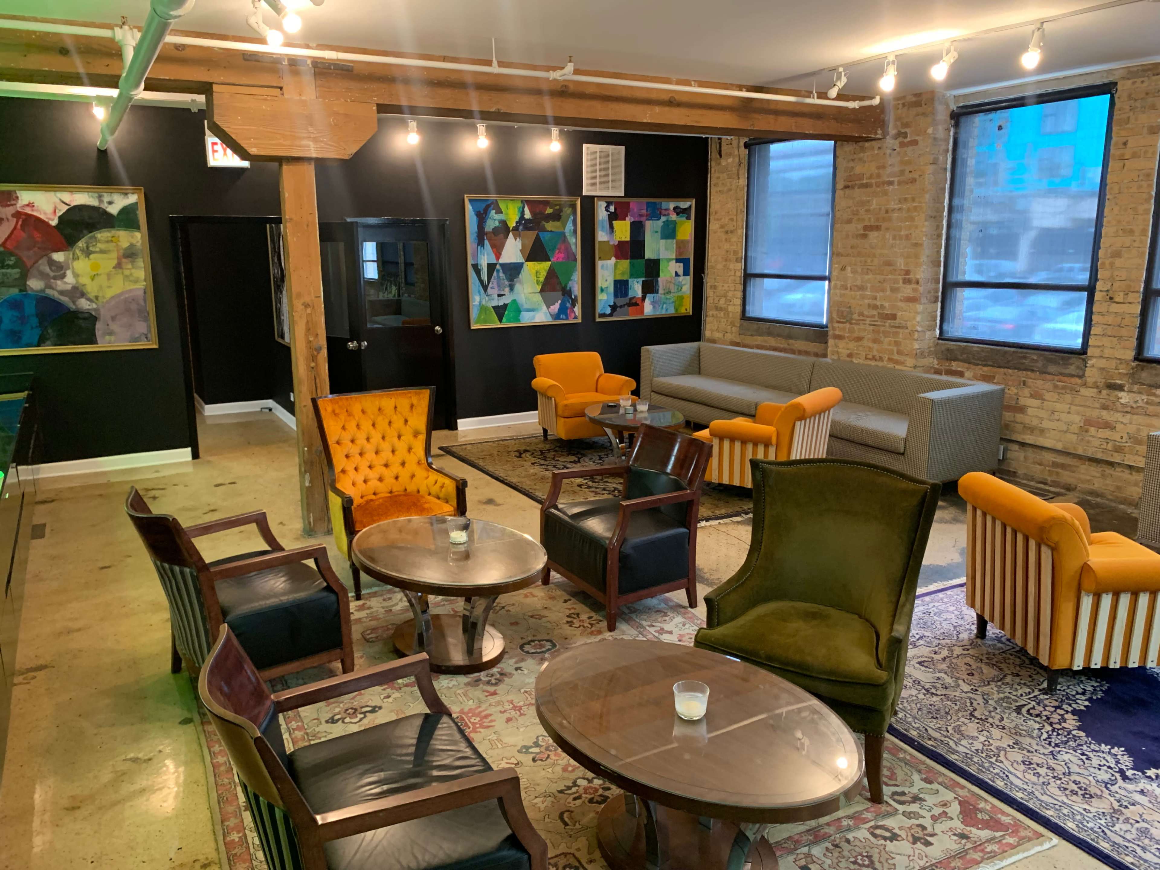 A modern lounge area with a mix of seating arrangements, including orange chairs, a gray sofa, and various tables on a patterned rug.