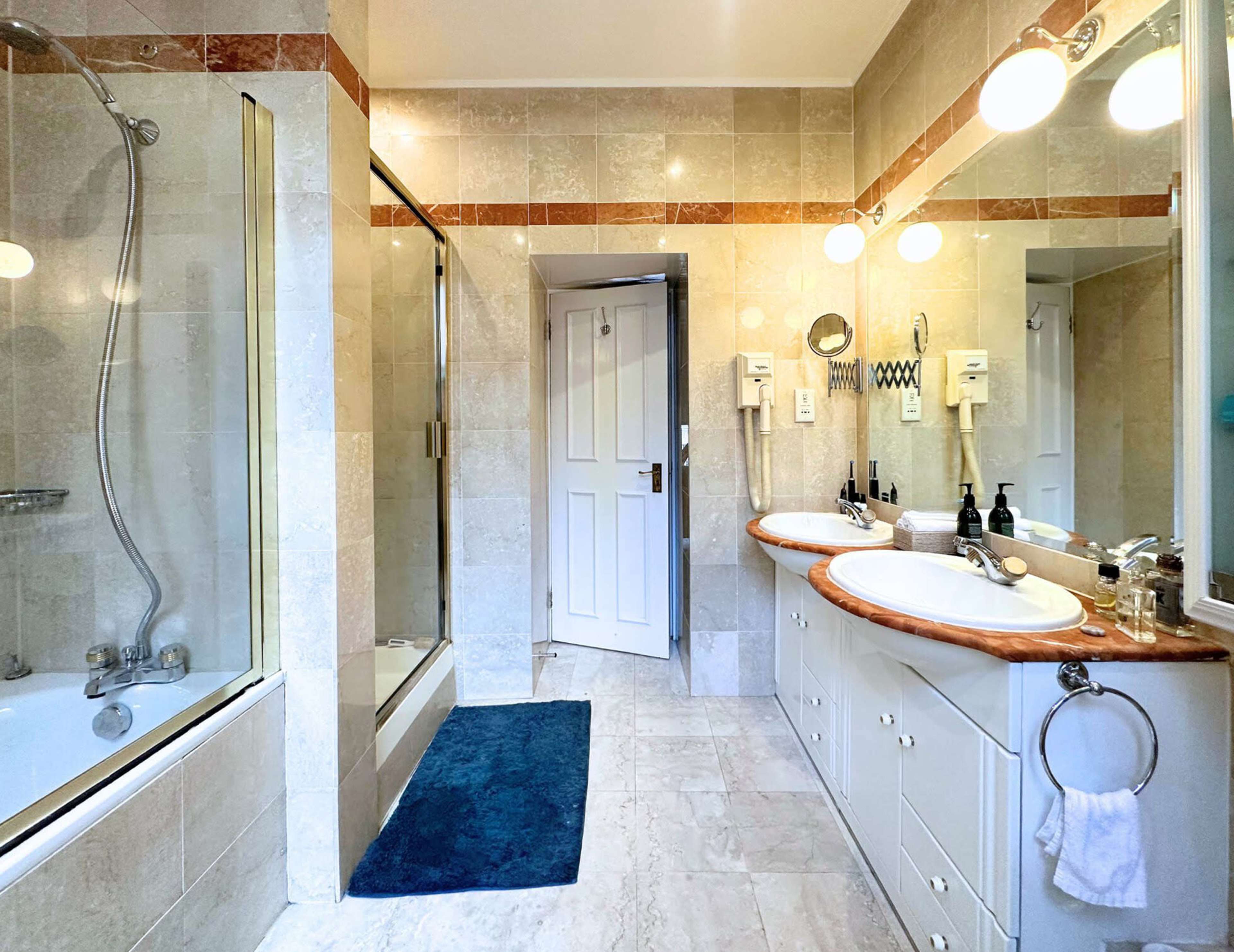 A spacious bathroom featuring a double vanity with sinks, a glass shower enclosure, a bathtub, and beige stone tiles.