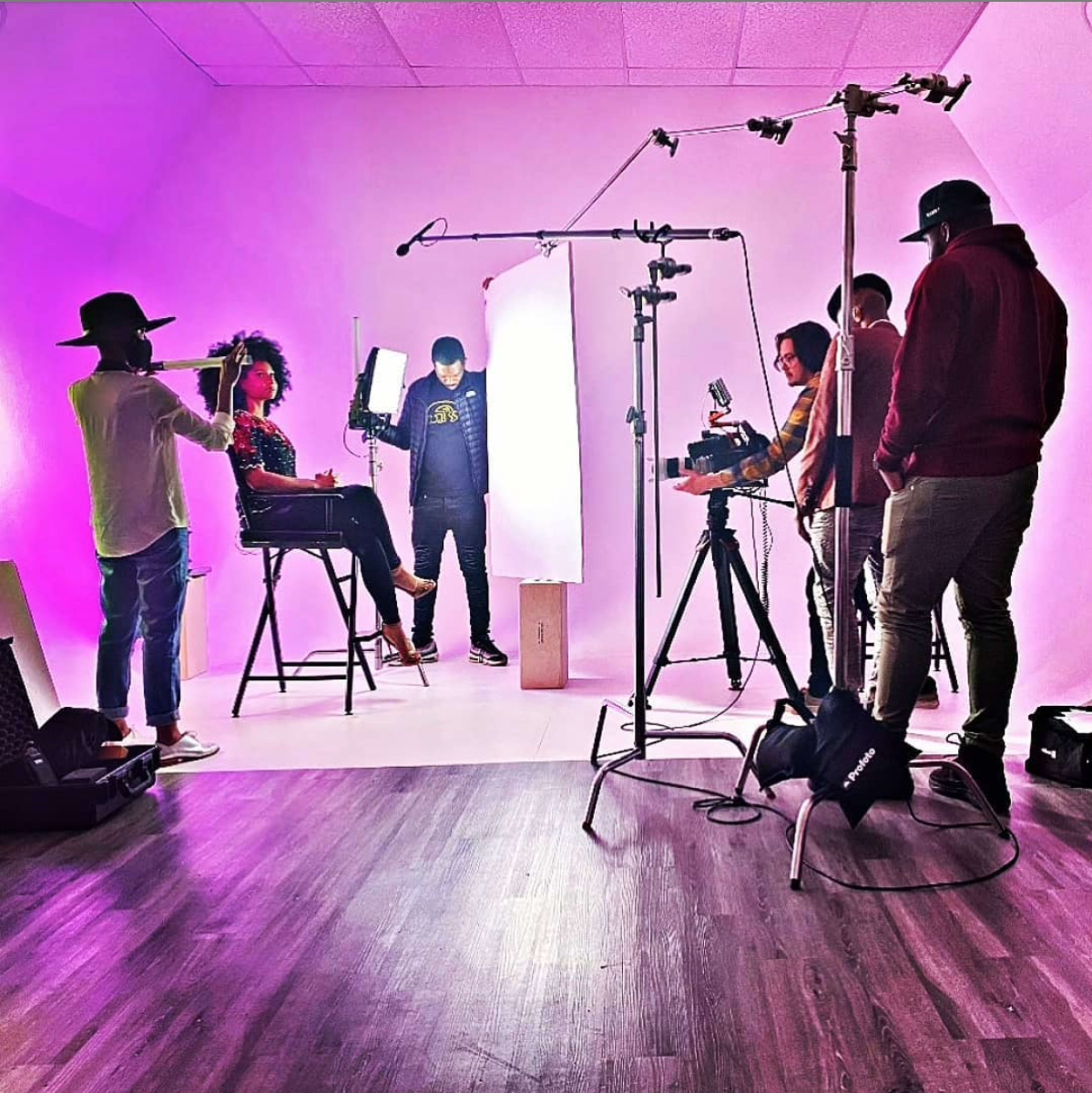 A group of six individuals is set up in a studio, with one person seated on a stool, while others operate cameras and lighting equipment in front of a brightly lit background.