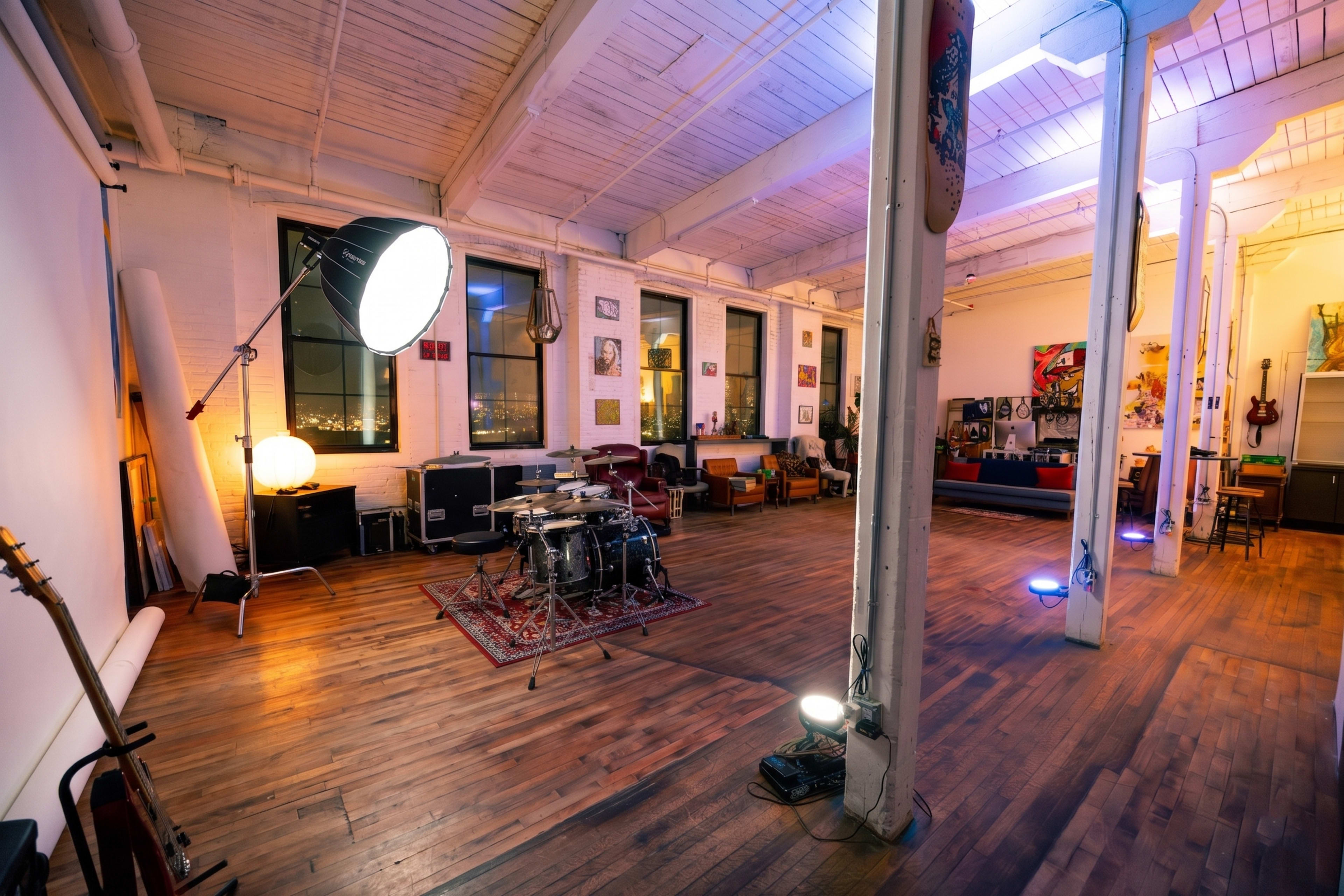 The image shows a spacious, well-lit studio featuring a drum set in the center, surrounded by seating areas and various artwork on the walls.