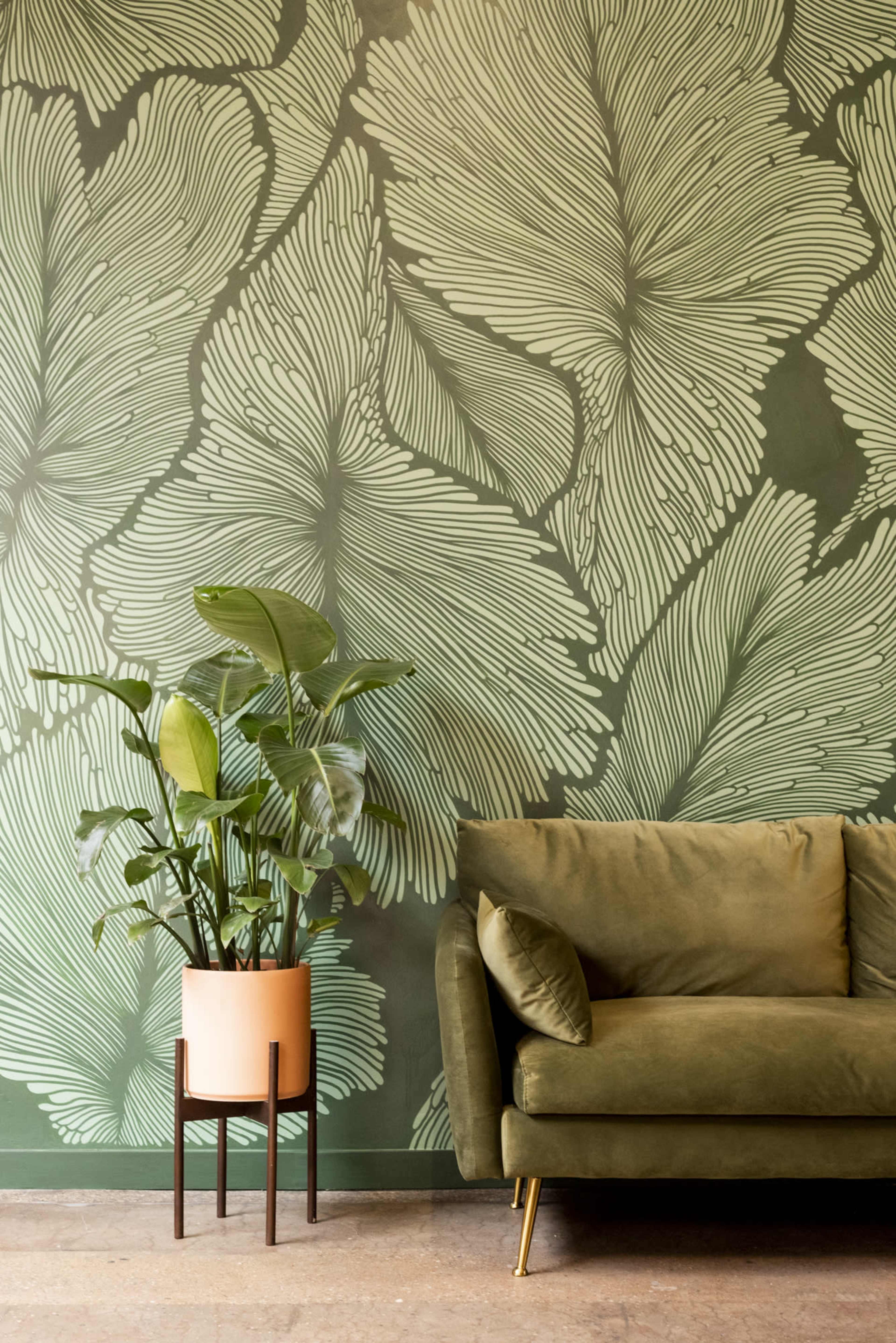A green sofa sits next to a potted plant against a wall adorned with a leafy green mural.