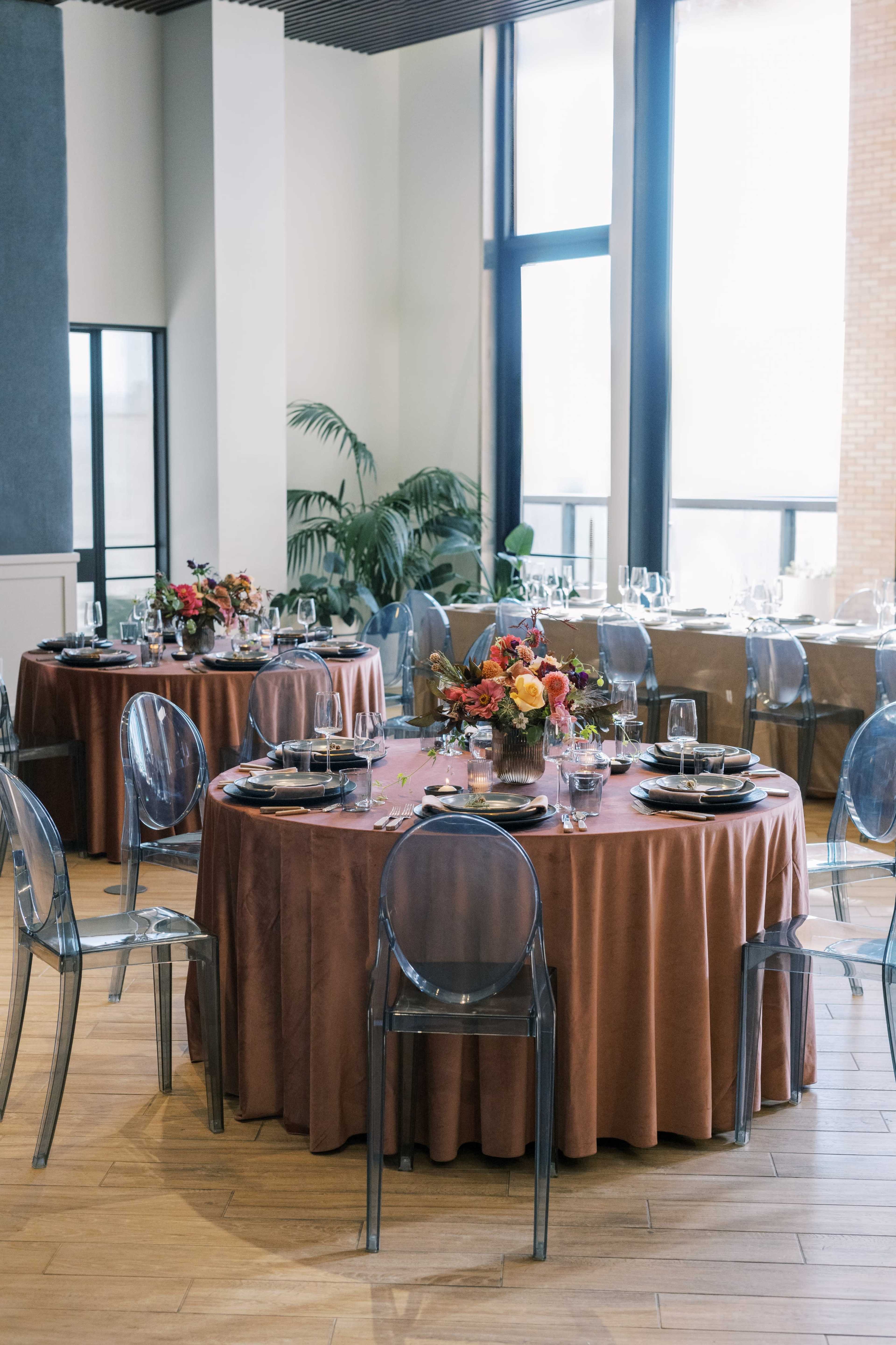 The image depicts a modern dining space with several round tables covered in brown tablecloths, adorned with floral centerpieces and clear ghost chairs, bathed in natural light from large windows.