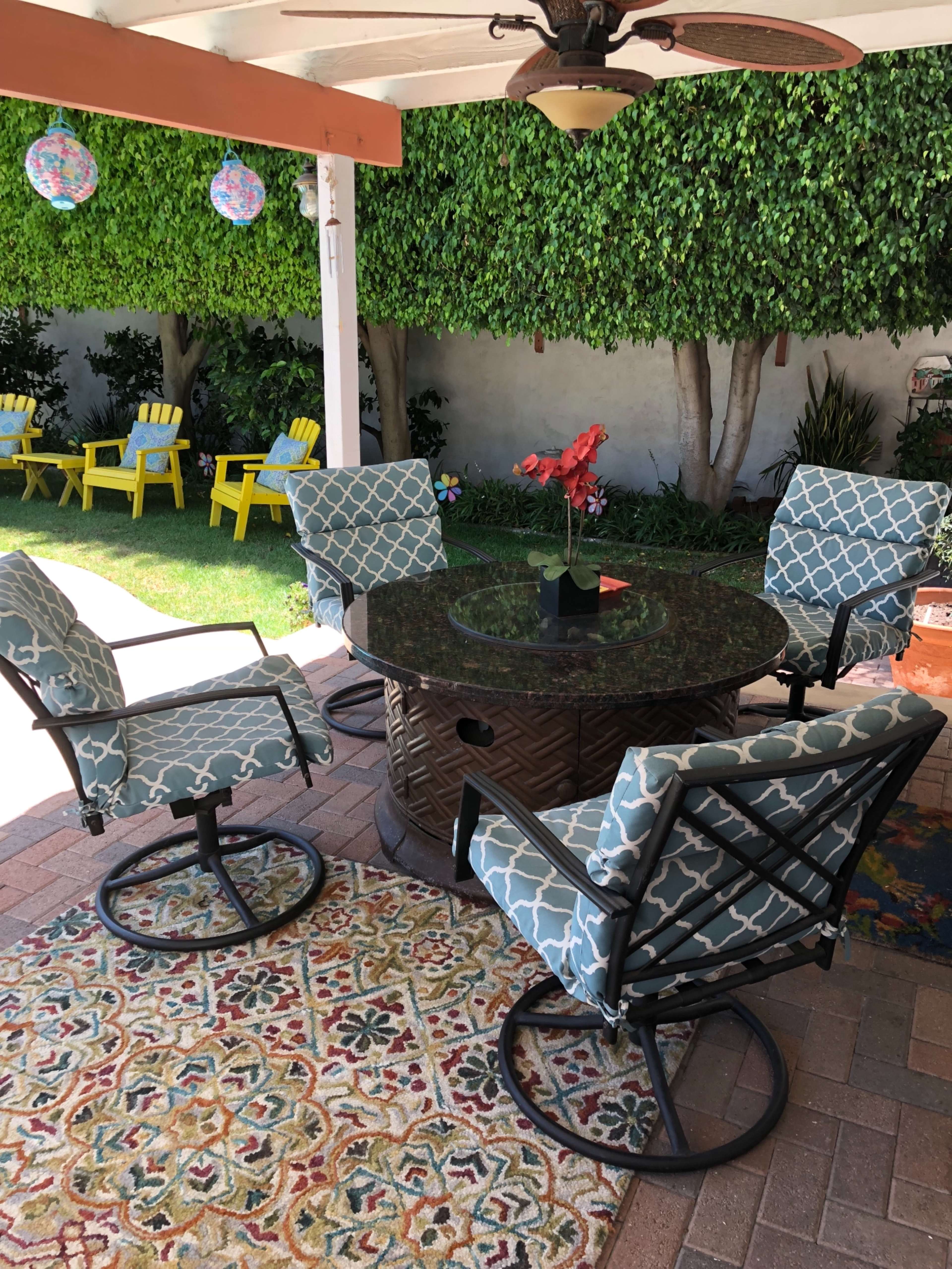 A round table with four patterned chairs sits under a covered patio surrounded by green hedges and a colorful outdoor rug.