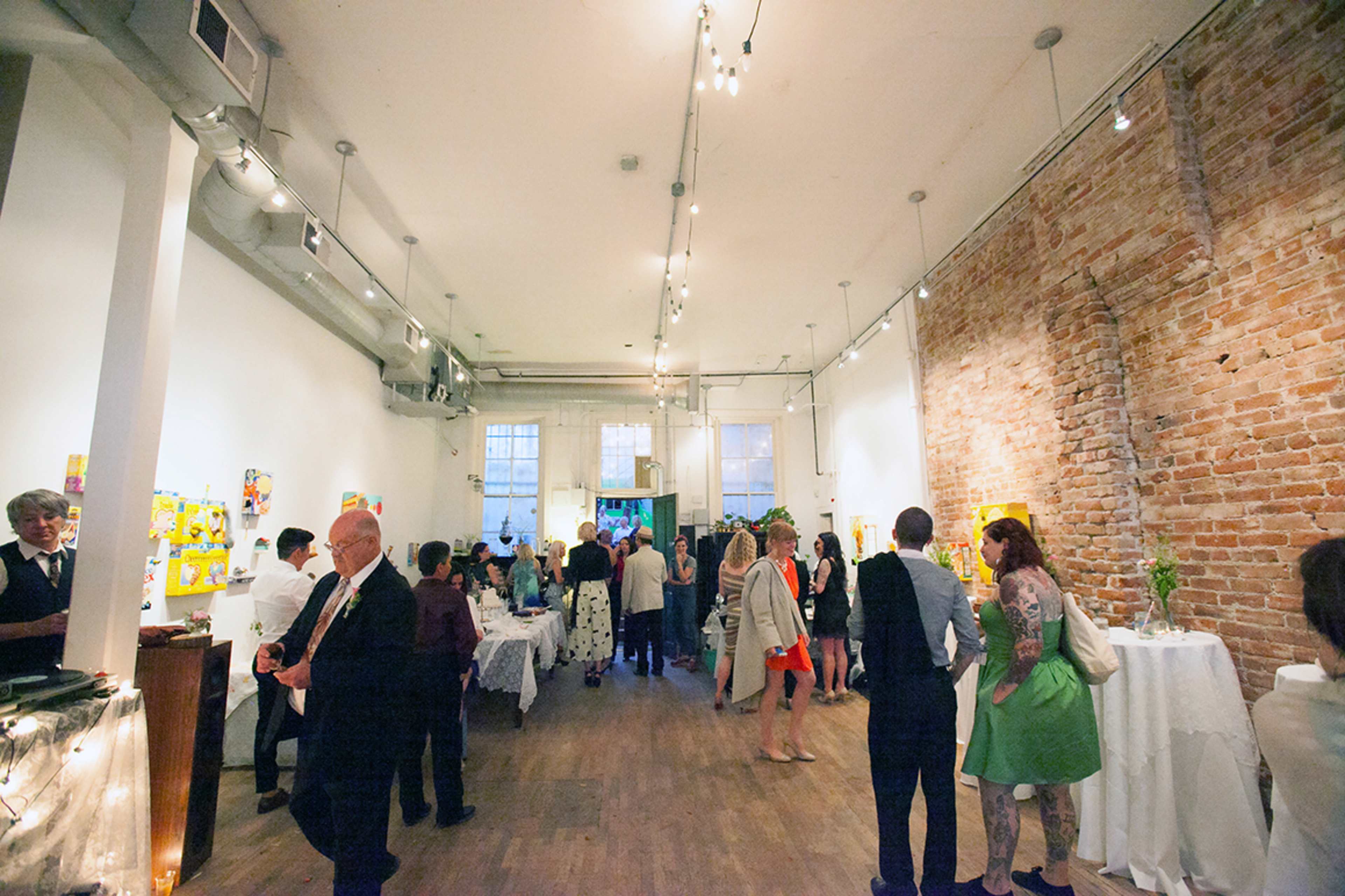 A crowded art gallery features guests mingling among colorful paintings on the walls, with white tablecloths and decorations.