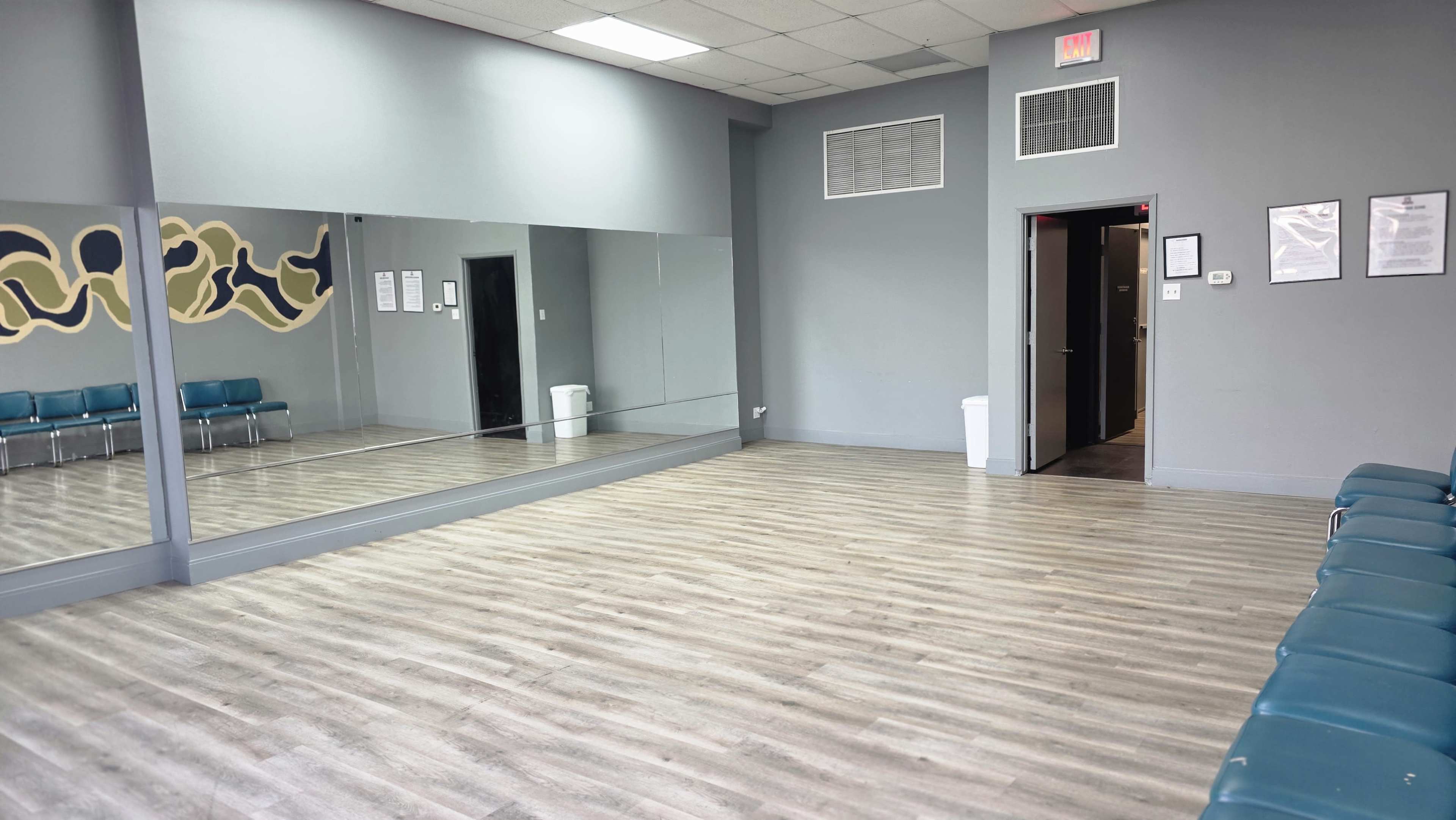 The image shows a spacious room with wooden floors, a large mirror on one wall, and a line of teal chairs against another wall.