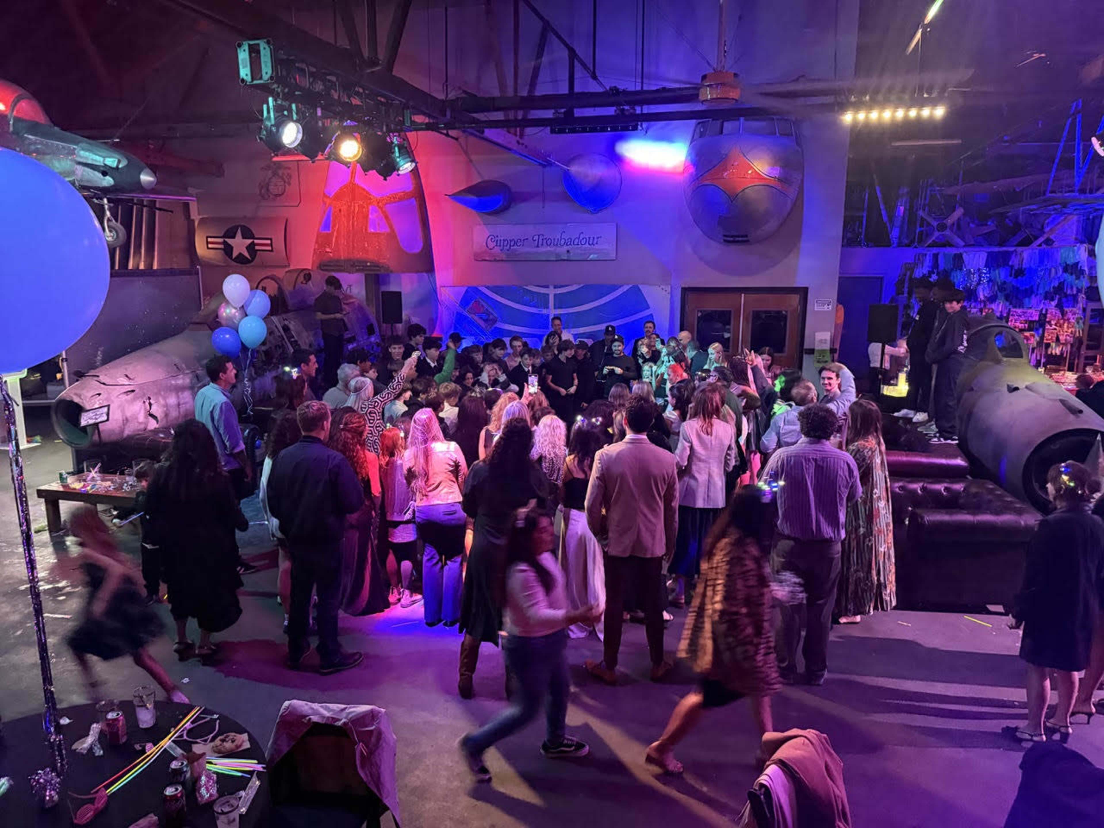 A large crowd of people gathers in a dimly lit event space decorated with aircraft memorabilia and colorful lights.