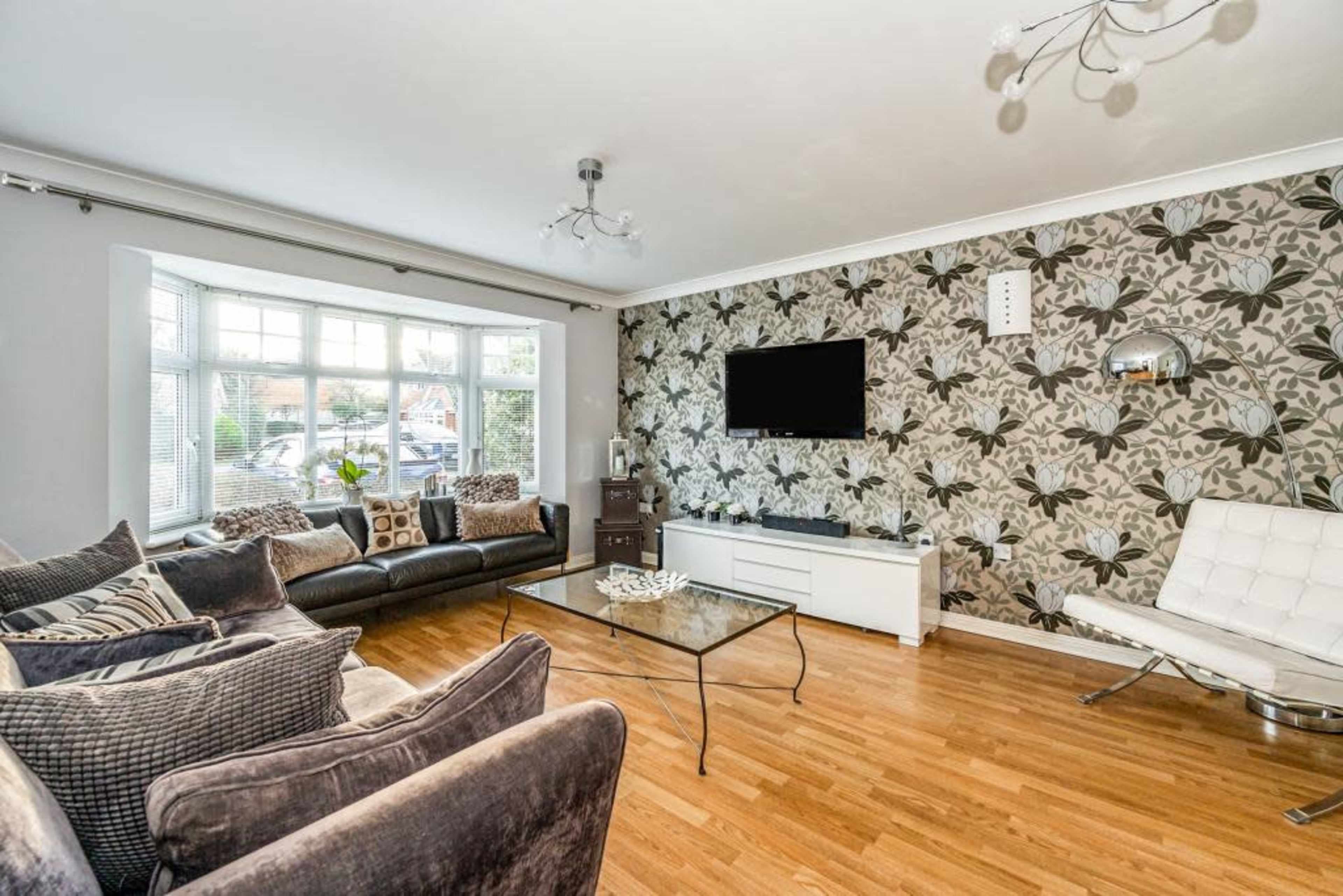 A modern living room features a large window, a sectional sofa, a glass coffee table, and a wall adorned with floral wallpaper.