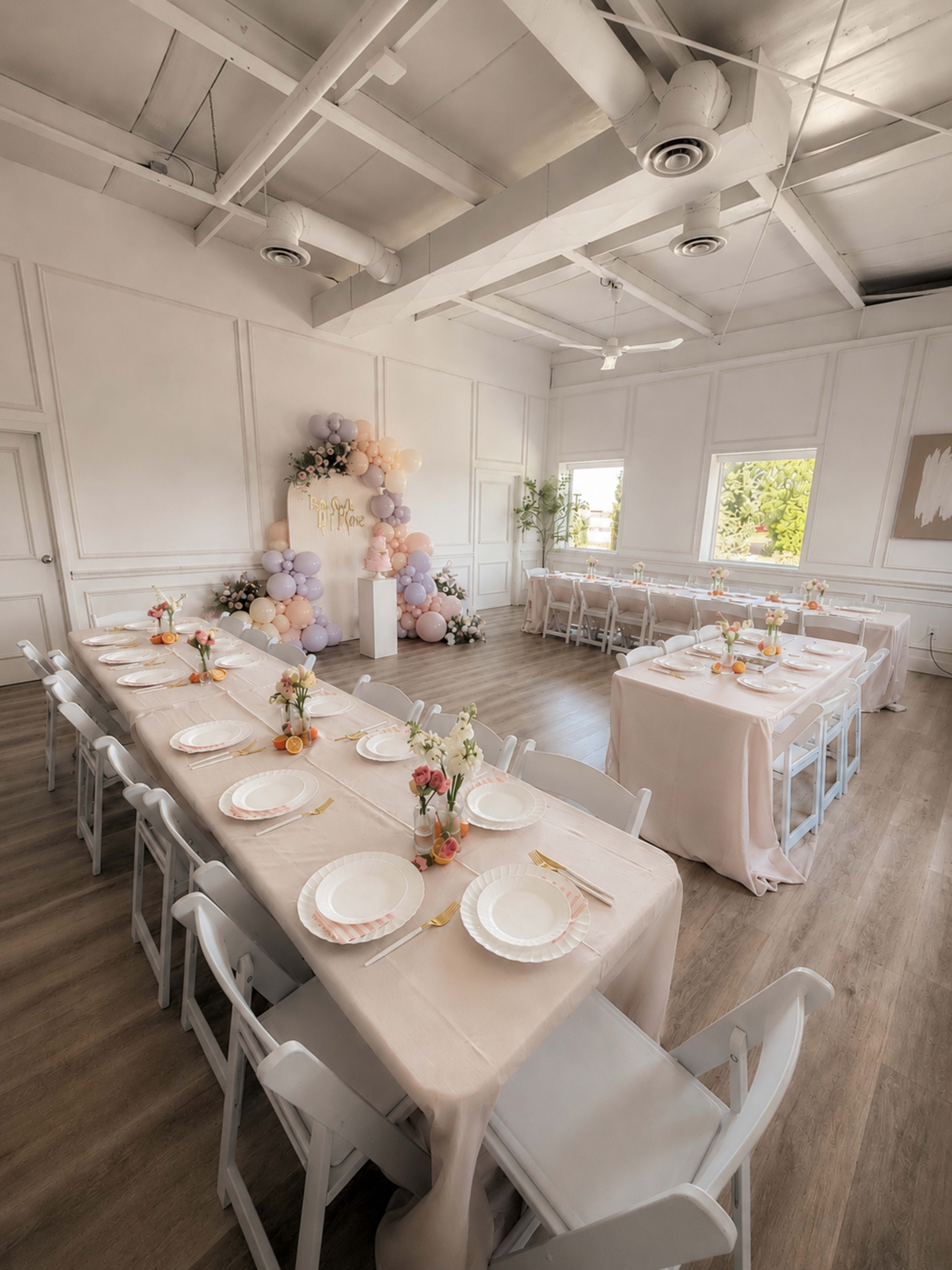 A large, elegantly decorated event space features multiple long tables set with floral centerpieces and pastel-colored tablecloths, alongside a backdrop adorned with balloons and floral arrangements.