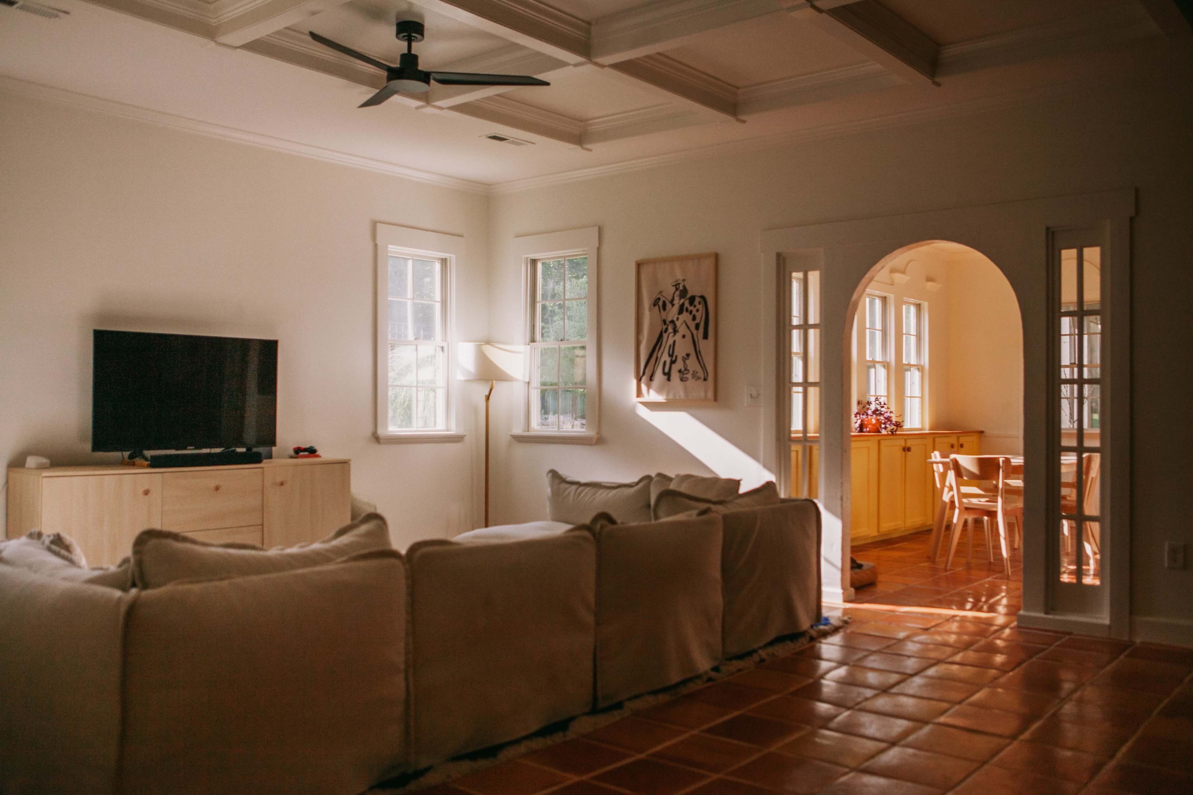 A spacious living room features a large sectional couch, a television, and natural light flowing through multiple windows, with an adjoining area visible through an archway.