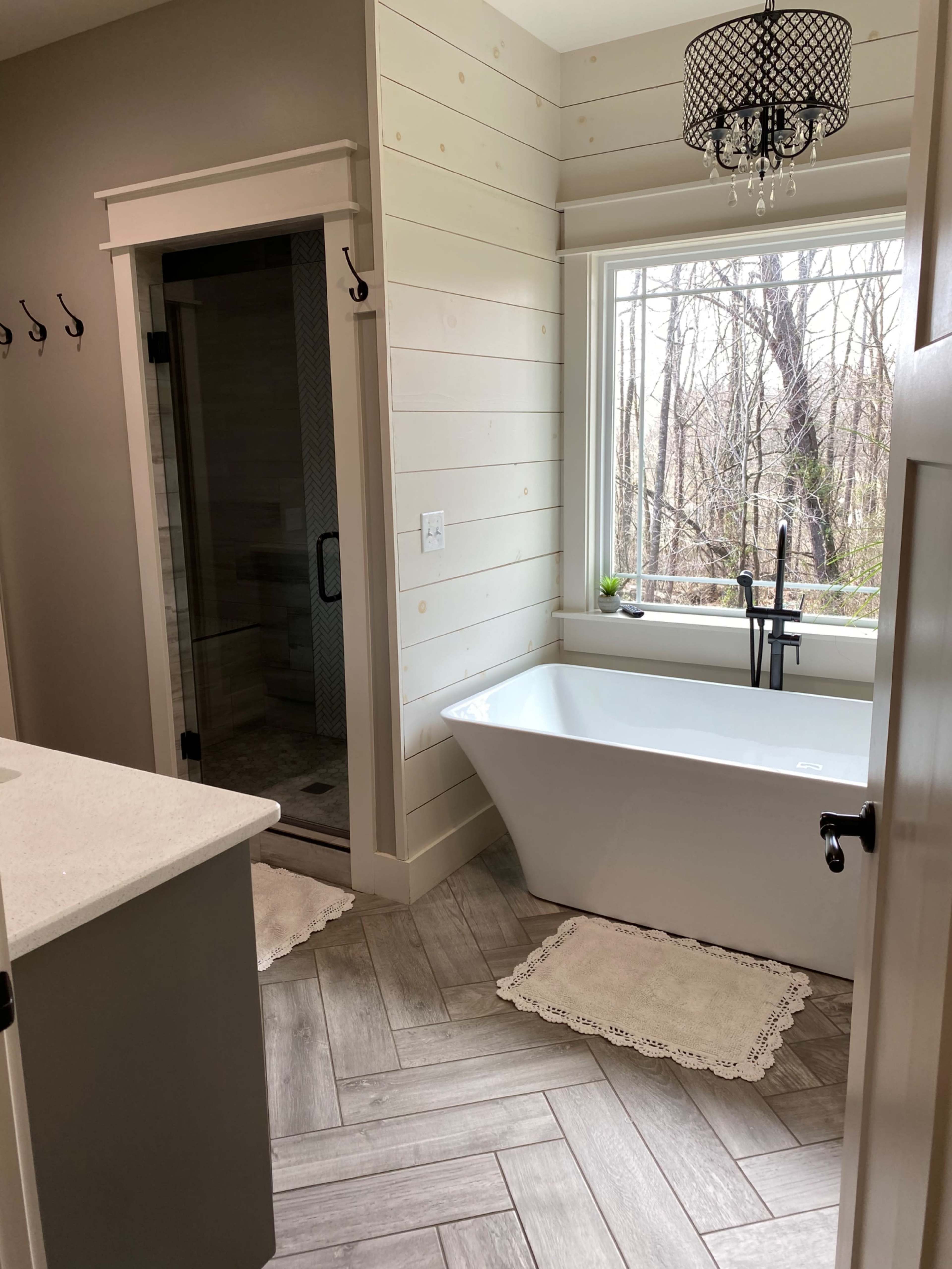 A modern bathroom featuring a freestanding bathtub, a glass shower, and wooden accent walls with a large window overlooking a wooded area.