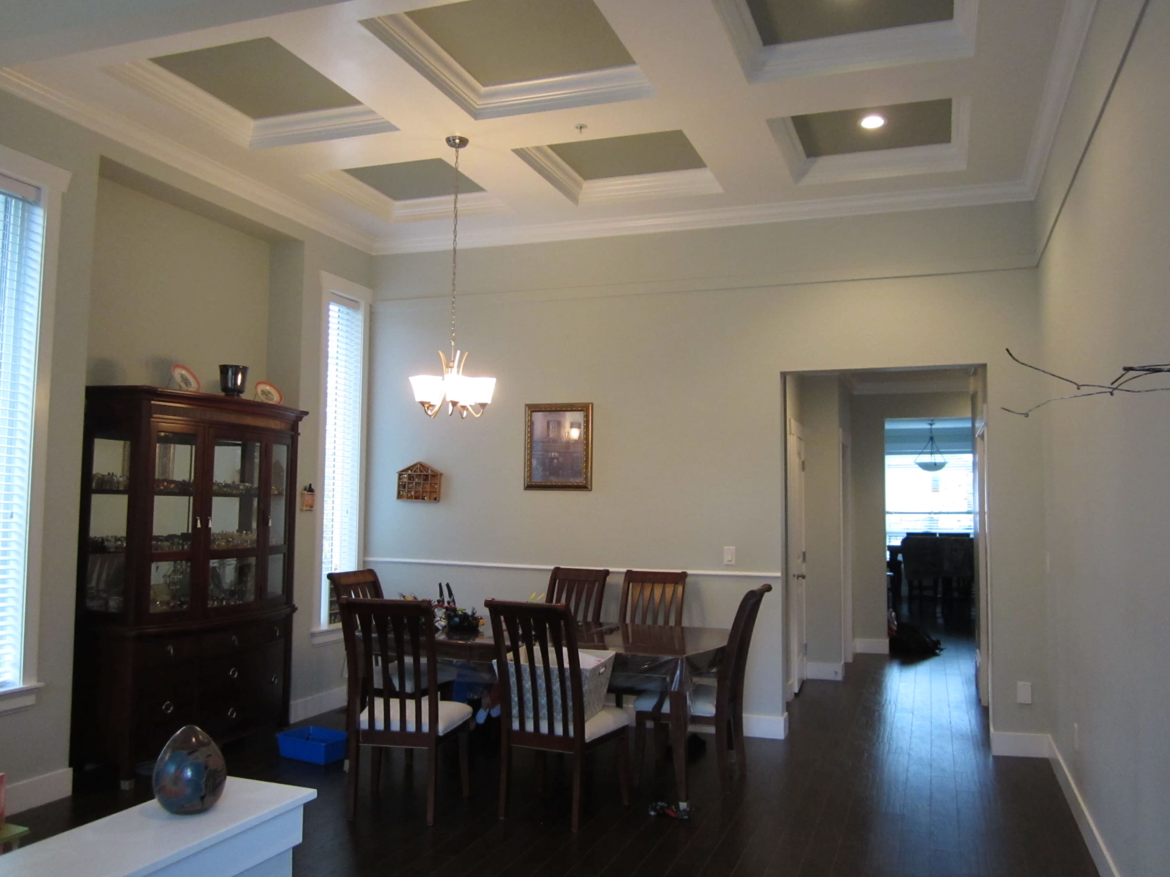 A dining room features a rectangular table surrounded by wooden chairs, with a coffered ceiling and a built-in cabinet against the wall.