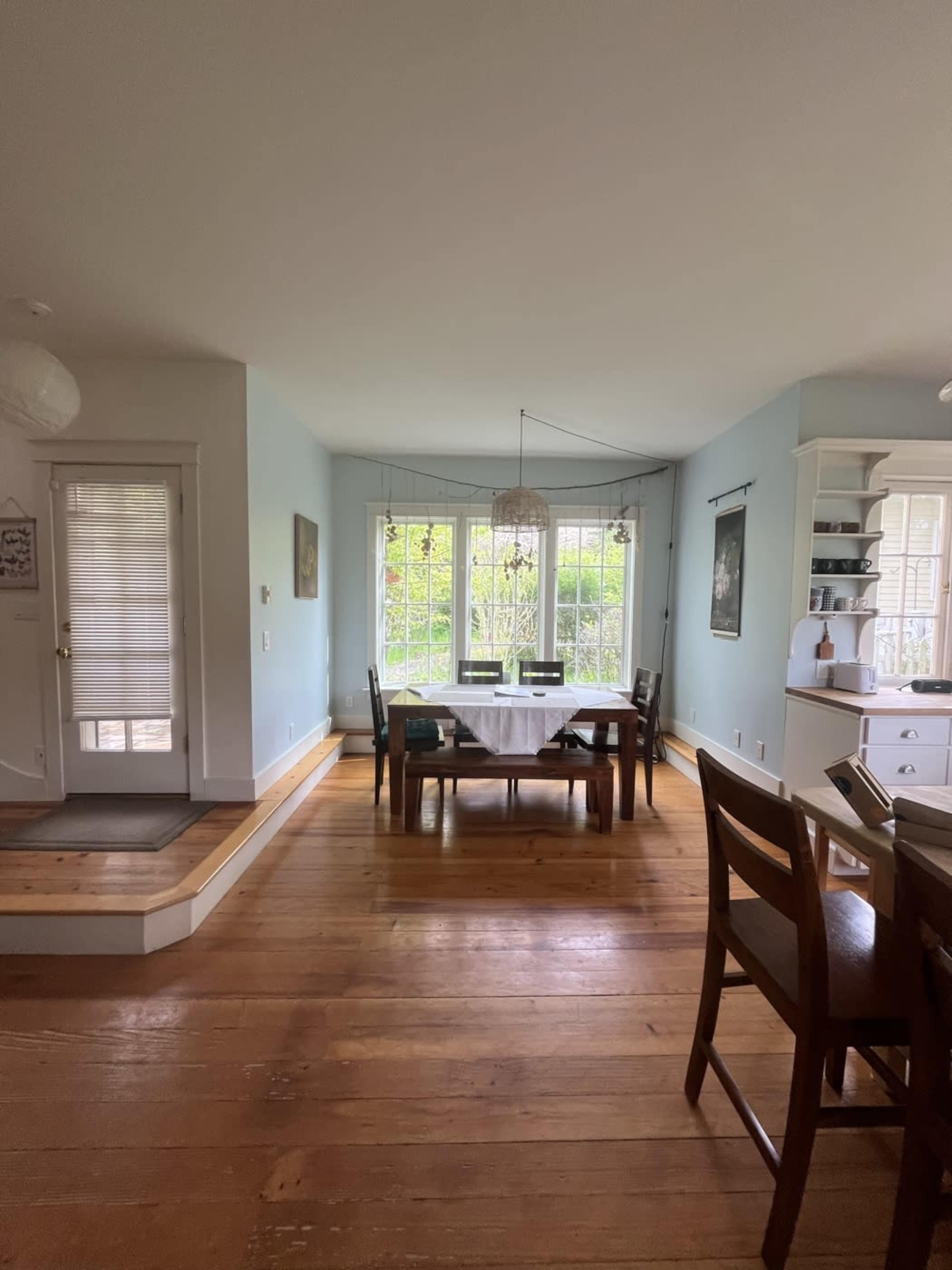 A dining area features a wooden table with chairs, large windows allowing natural light, and access to an adjacent room.