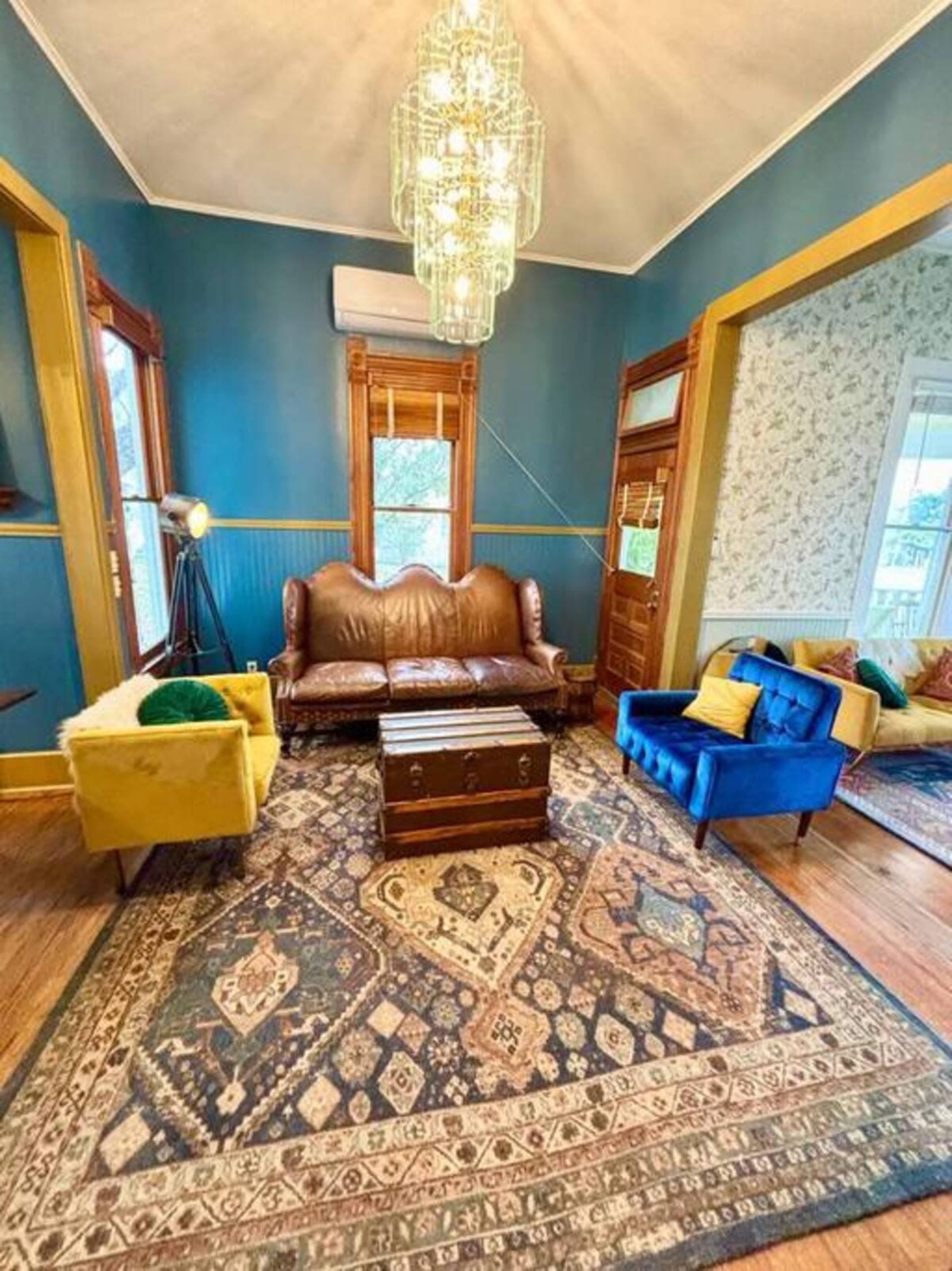 The image shows a living room with blue walls, a large patterned rug, a brown leather sofa, and a colorful blue armchair beside a vintage trunk.