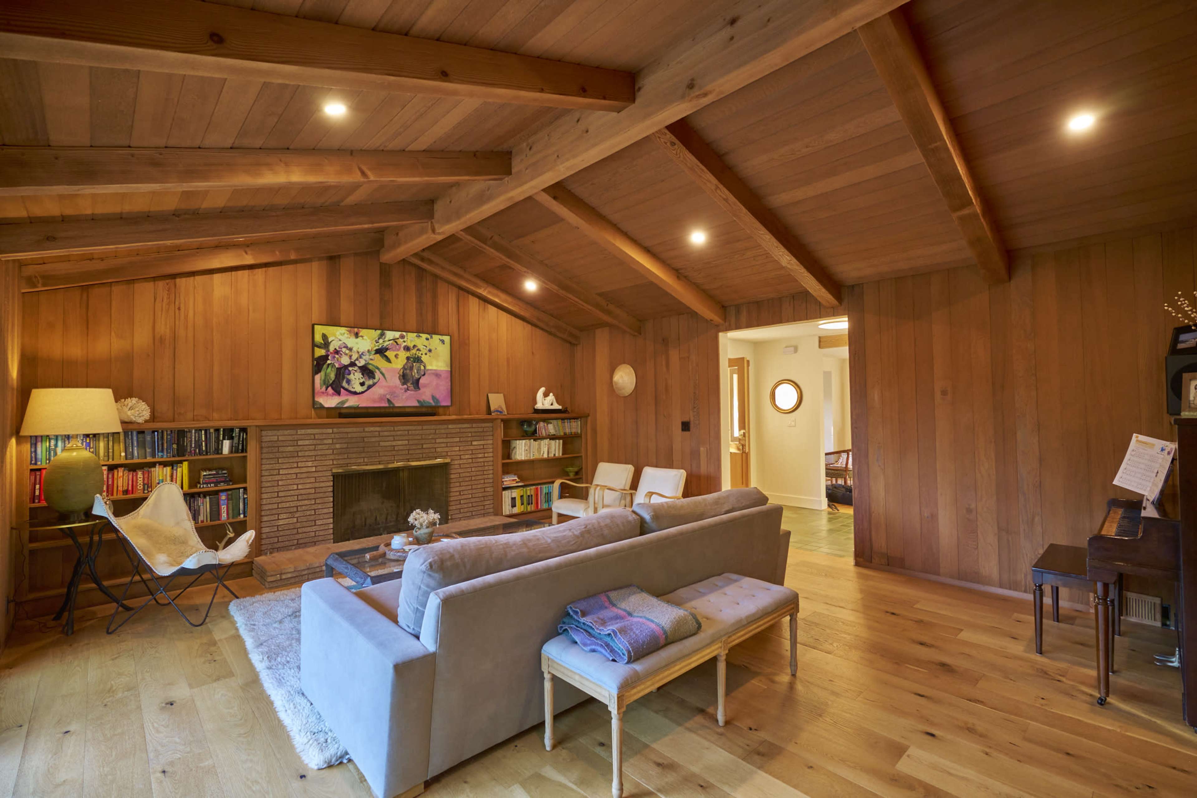 The living room features wooden walls and a ceiling with exposed beams, a brick fireplace, a light gray sofa, and a bookshelf, creating a cozy space.