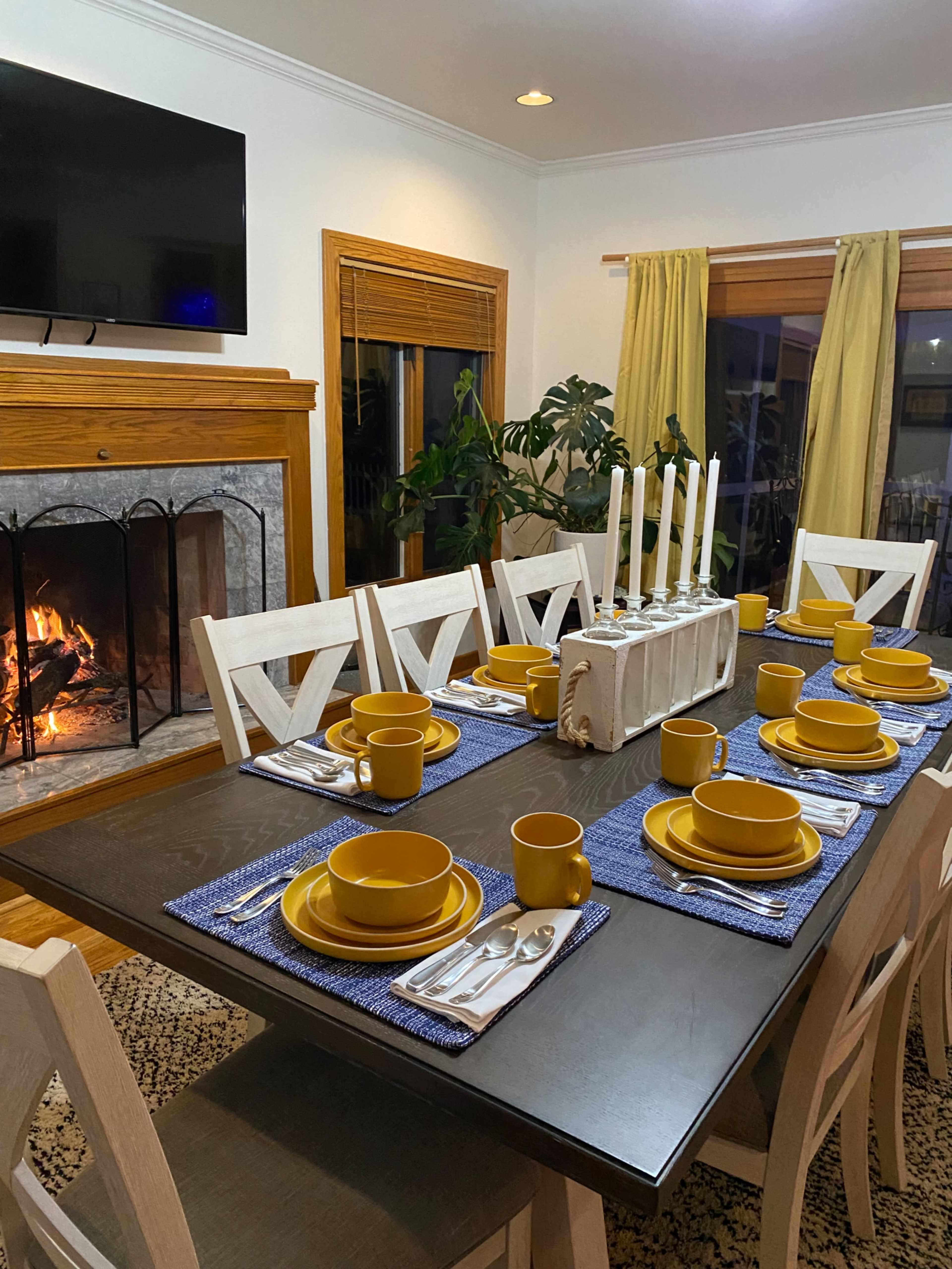 A dining table is set with yellow plates and cups on blue placemats, while a fireplace burns in the background.