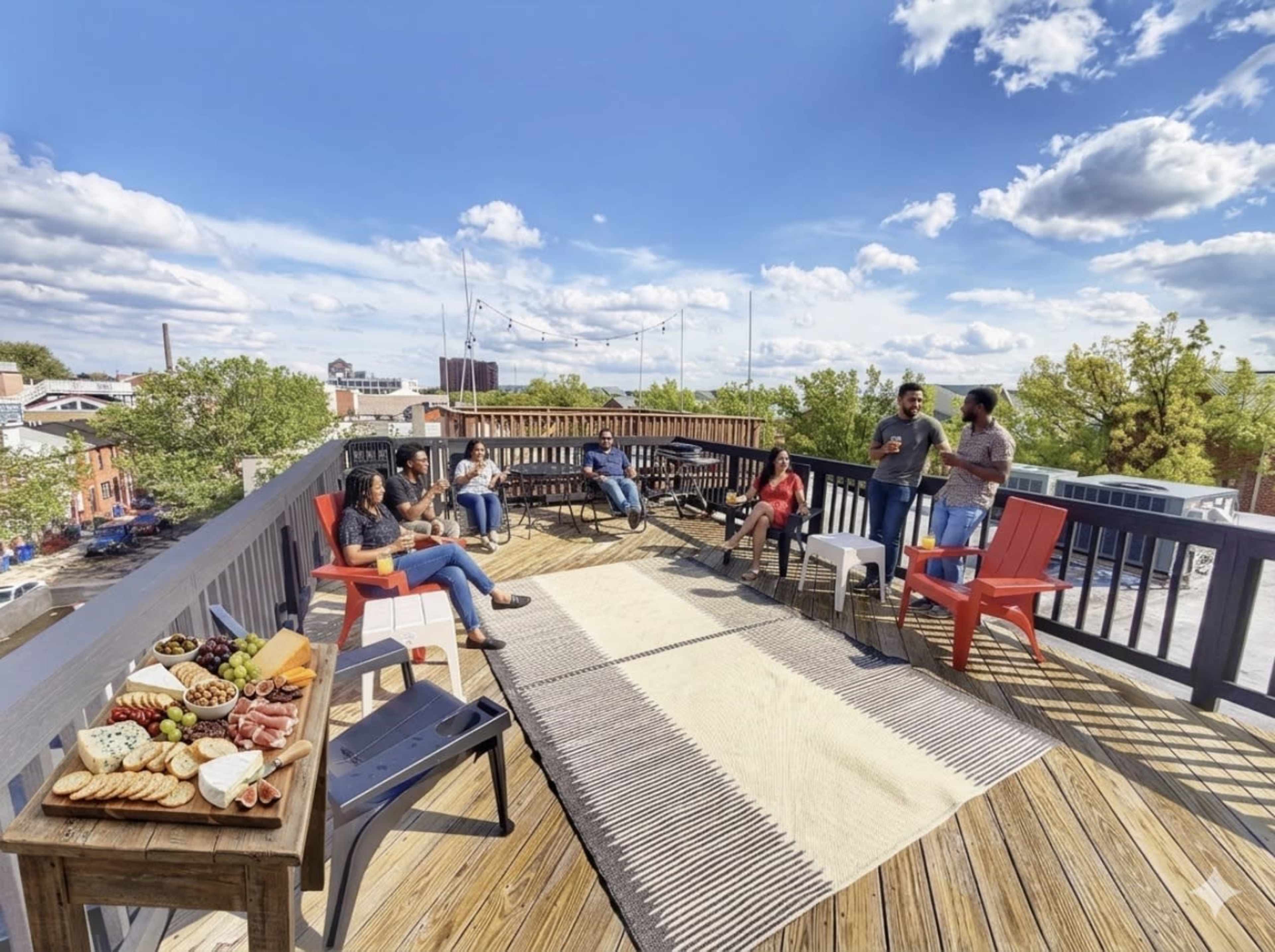 A group of people gathers on a rooftop deck with various snacks and drinks, surrounded by greenery and blue skies.